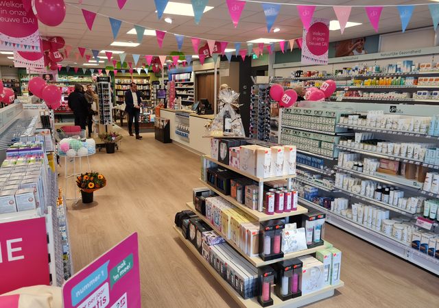 Een helder verlicht apotheekinterieur met schappen met producten, roze en blauwe vlaggetjes, ballonnen en een paar rondsnuffelende klanten.