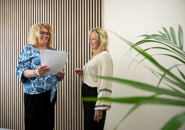 Twee vrouwen glimlachen en bespreken een document in een modern kantoor met houten lambrisering en een plant op de voorgrond.