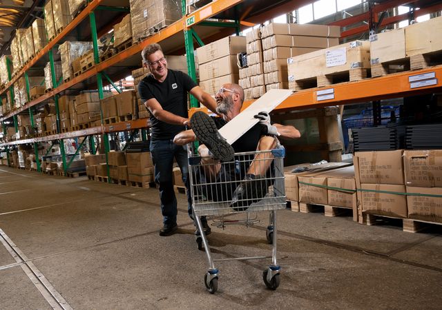 Twee mensen in een magazijn: de één duwt een winkelwagentje terwijl de ander binnen zit, omringd door planken met dozen en pakjes.