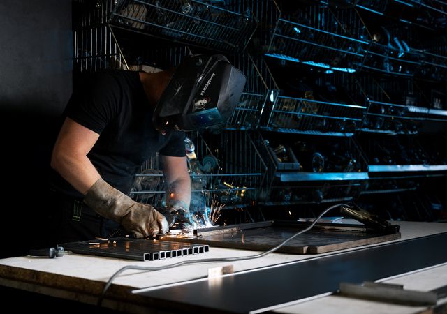 Persoon die metaal las op een werkbank, beschermende kleding draagt, met planken met gereedschap op de achtergrond. Er zijn vonken zichtbaar tijdens het lasproces.