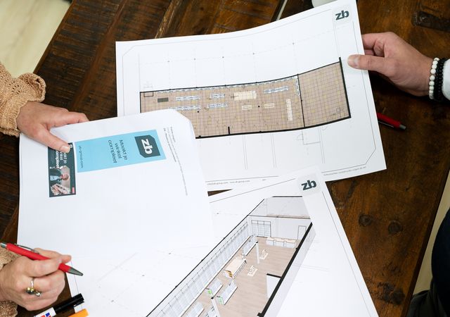 Twee mensen beoordelen architectonische plannen op een houten tafel, met vloerindelingen en ontwerpdetails.