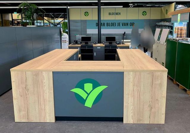 Houten U-vormige toonbank met groen logo in een winkel, met kassa's en een bord "BLOEMEN" erboven.