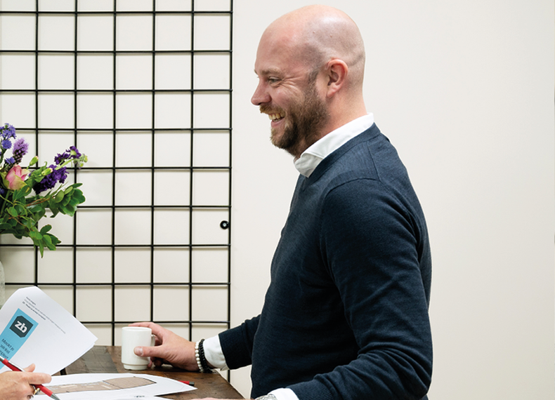 Een glimlachende man in een blauwe trui houdt een koffiekopje vast en staat bij een tafel met documenten, naast een rastermuur met bloemen op de achtergrond.