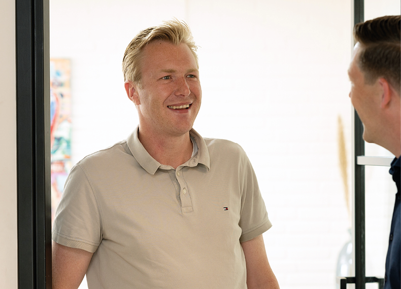 Een glimlachende man in een beige poloshirt staat binnen en is in gesprek met een andere persoon.