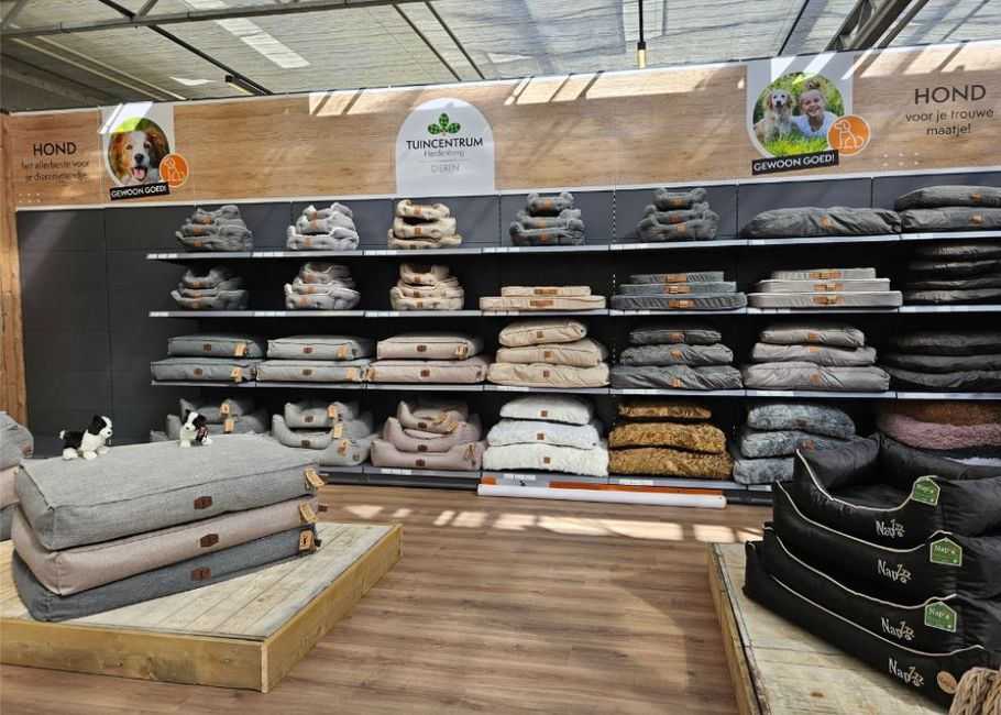 Dierenwinkelgedeelte met planken waarop verschillende hondenbedden in verschillende maten en kleuren staan, naast enkele knuffels.
