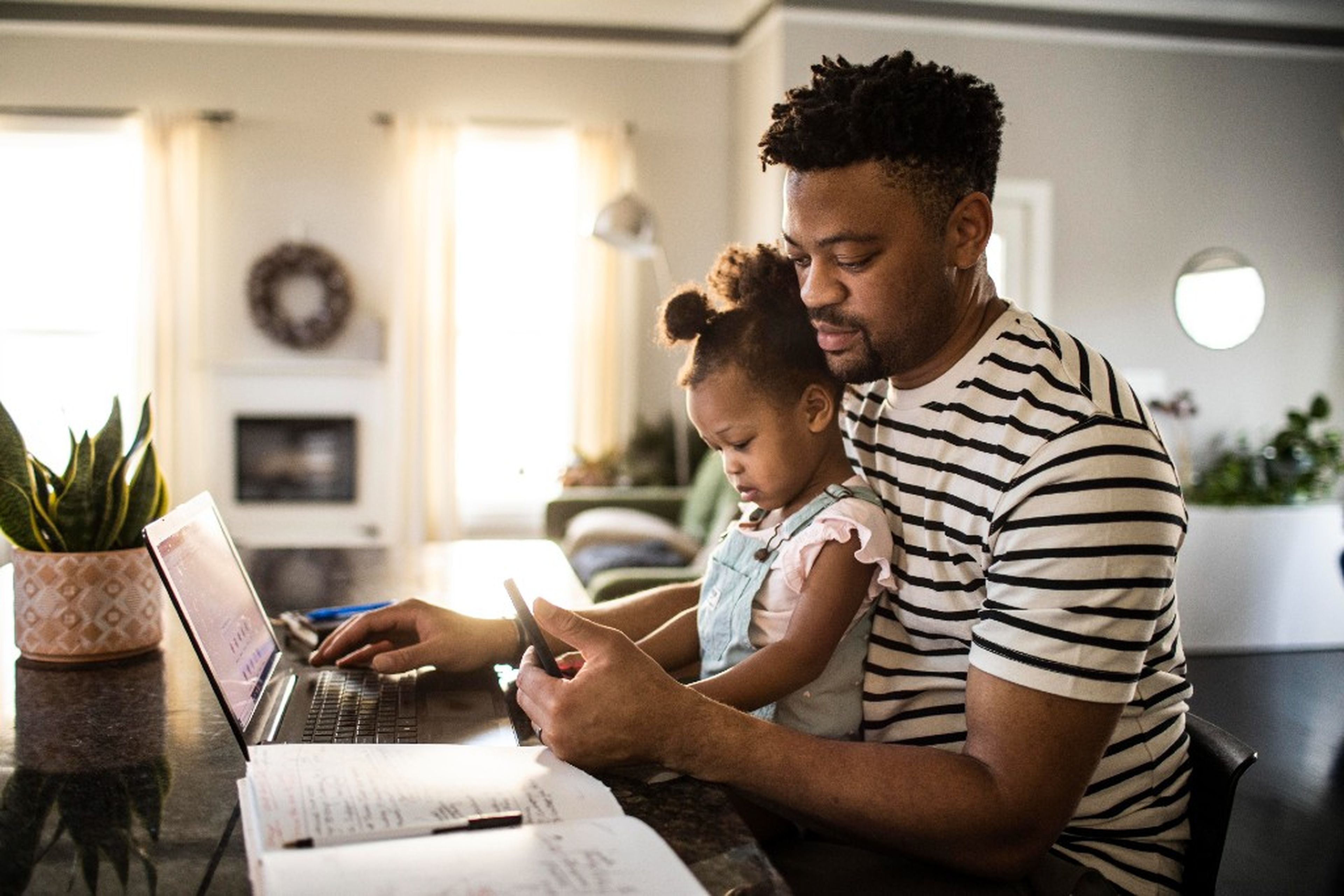 A man works on a laptop at a table in a cozy room, with a child on his lap