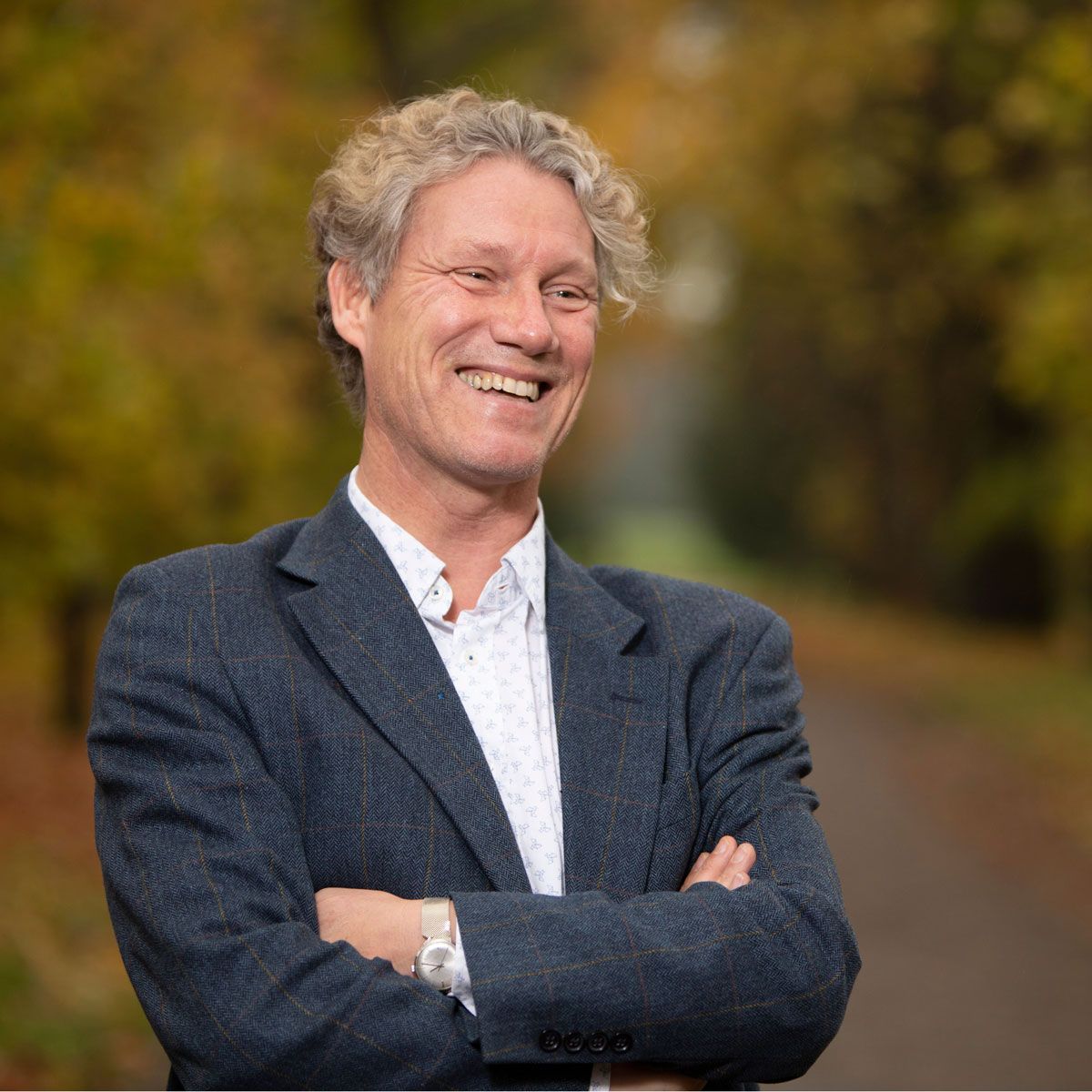 Smiling man with curly hair, arms crossed, wearing a blue blazer and white shirt outdoors, with a blurred autumnal background.