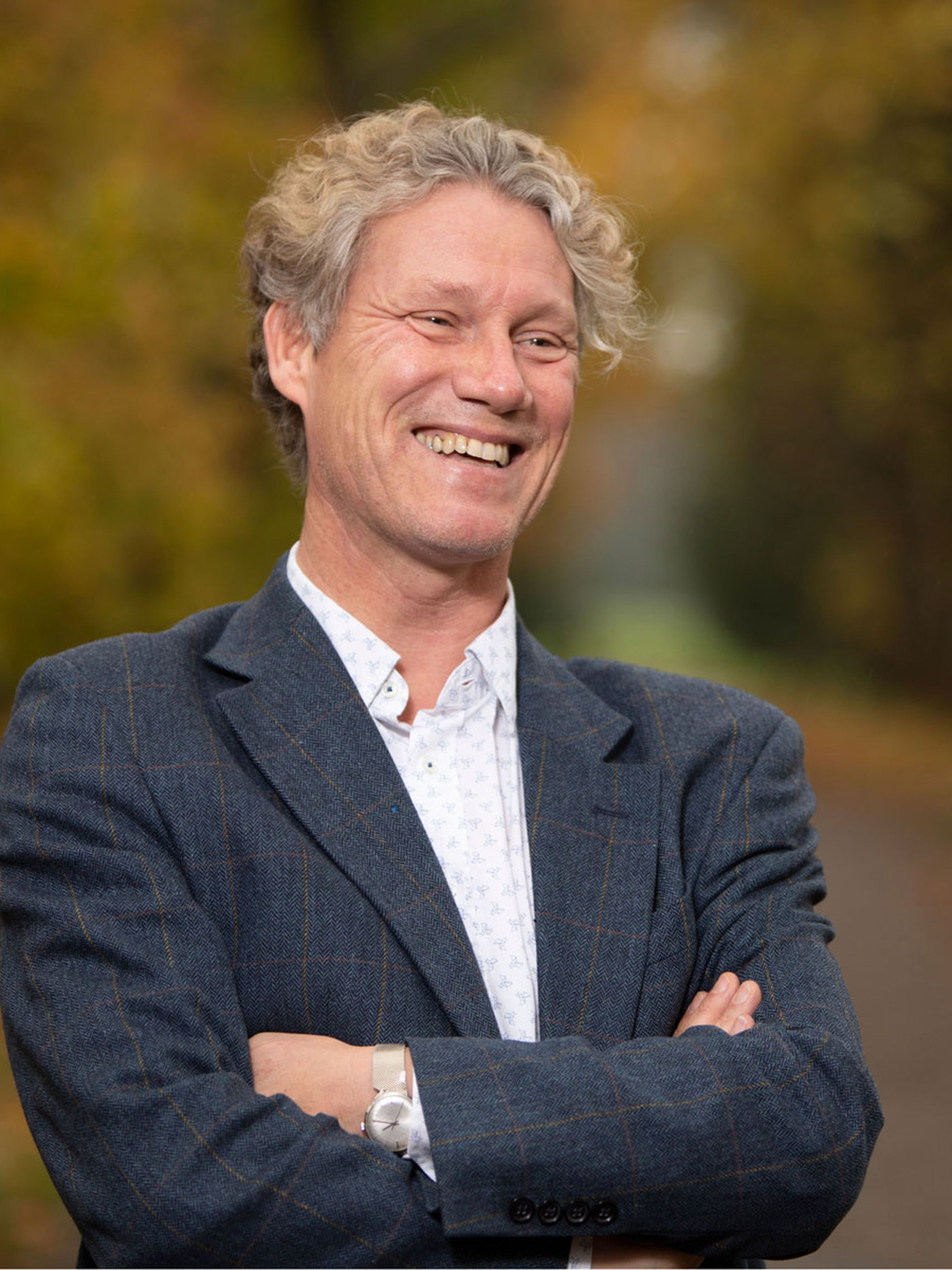 Smiling man with curly hair, arms crossed, wearing a blue blazer and white shirt outdoors, with a blurred autumnal background.