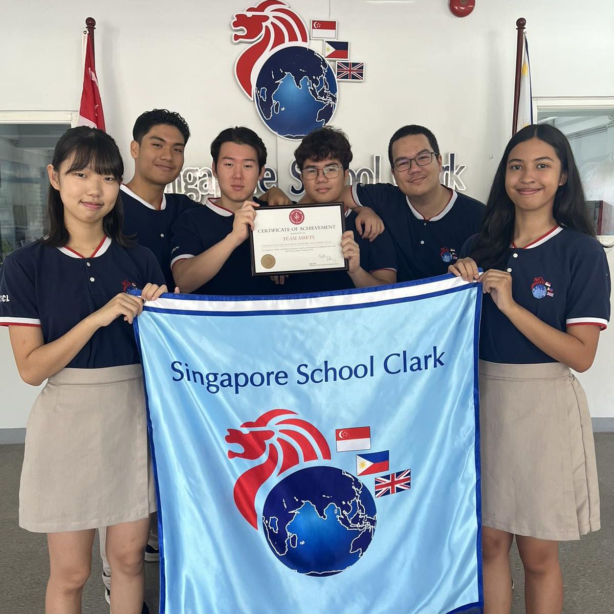 Group of students holding a "Certificate of Achievement" and a "Singapore School Clark" banner, standing in front of a logo.