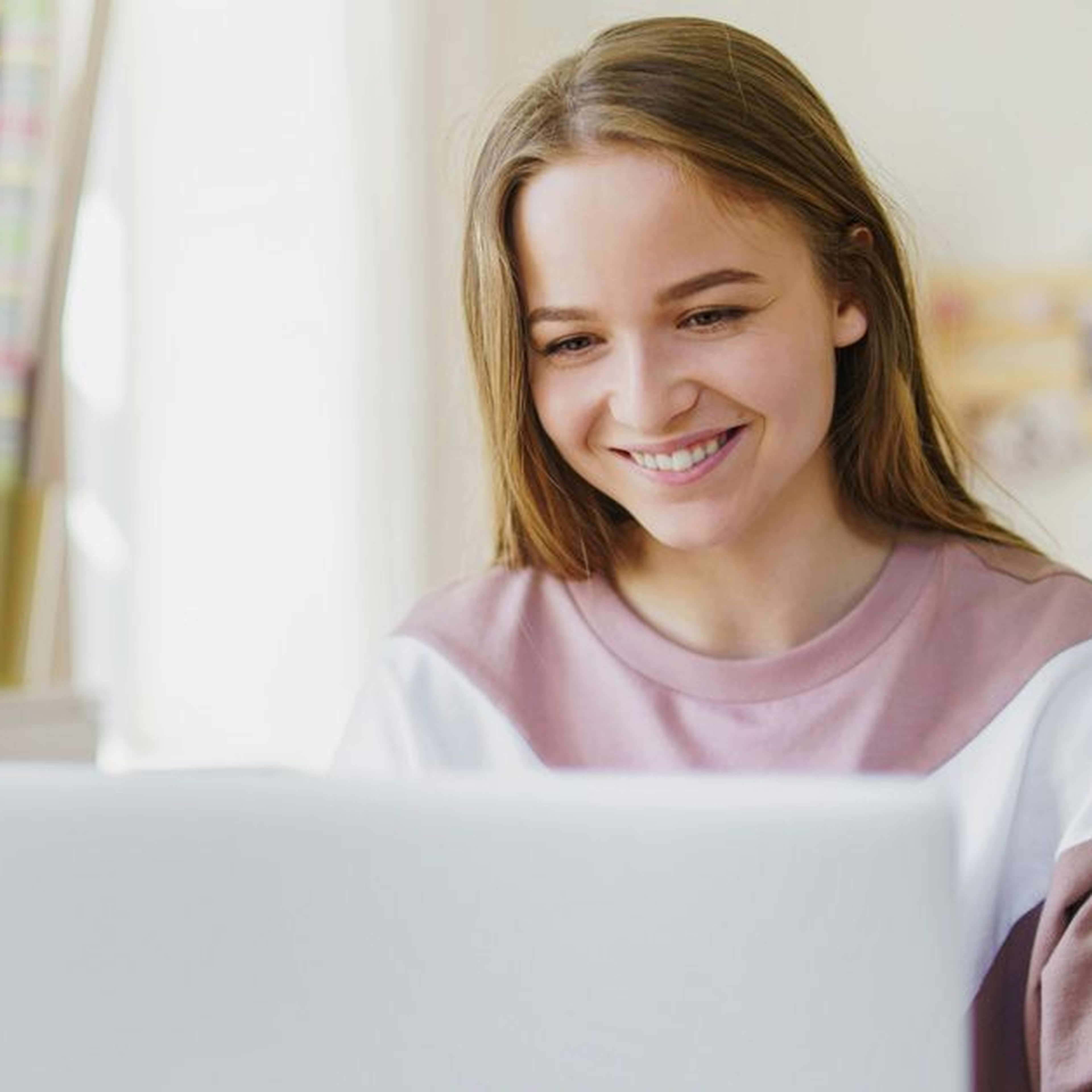 Smiling woman in a cozy room using a laptop, with a desk, lamp, and decorations in the background.