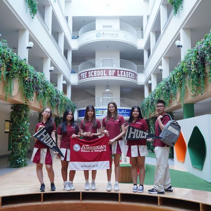 Six students in uniforms hold school banners inside a building with greenery and a "The School of Leaders" sign above.