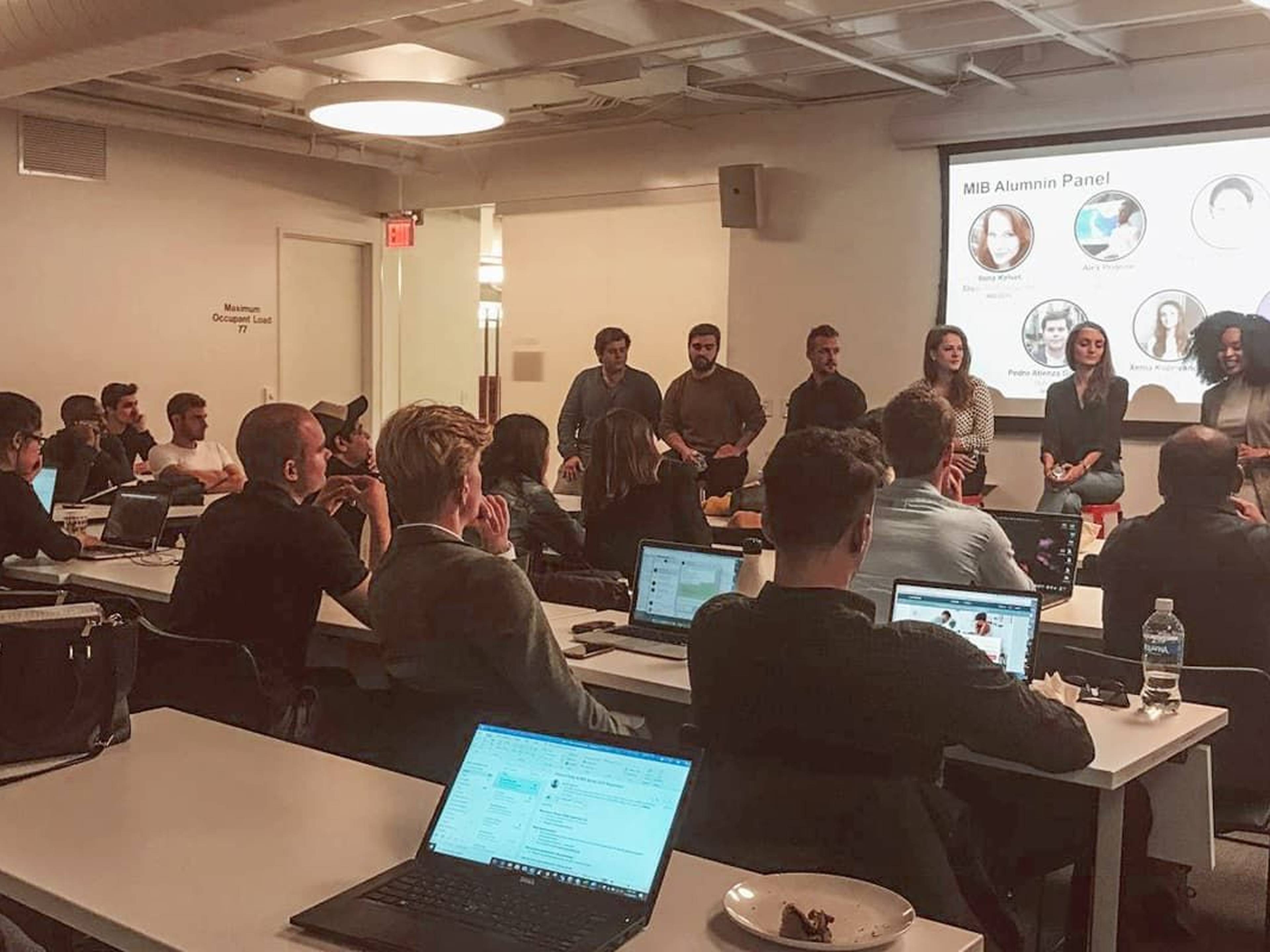 People in a classroom listen to a panel presentation with laptops open on desks.