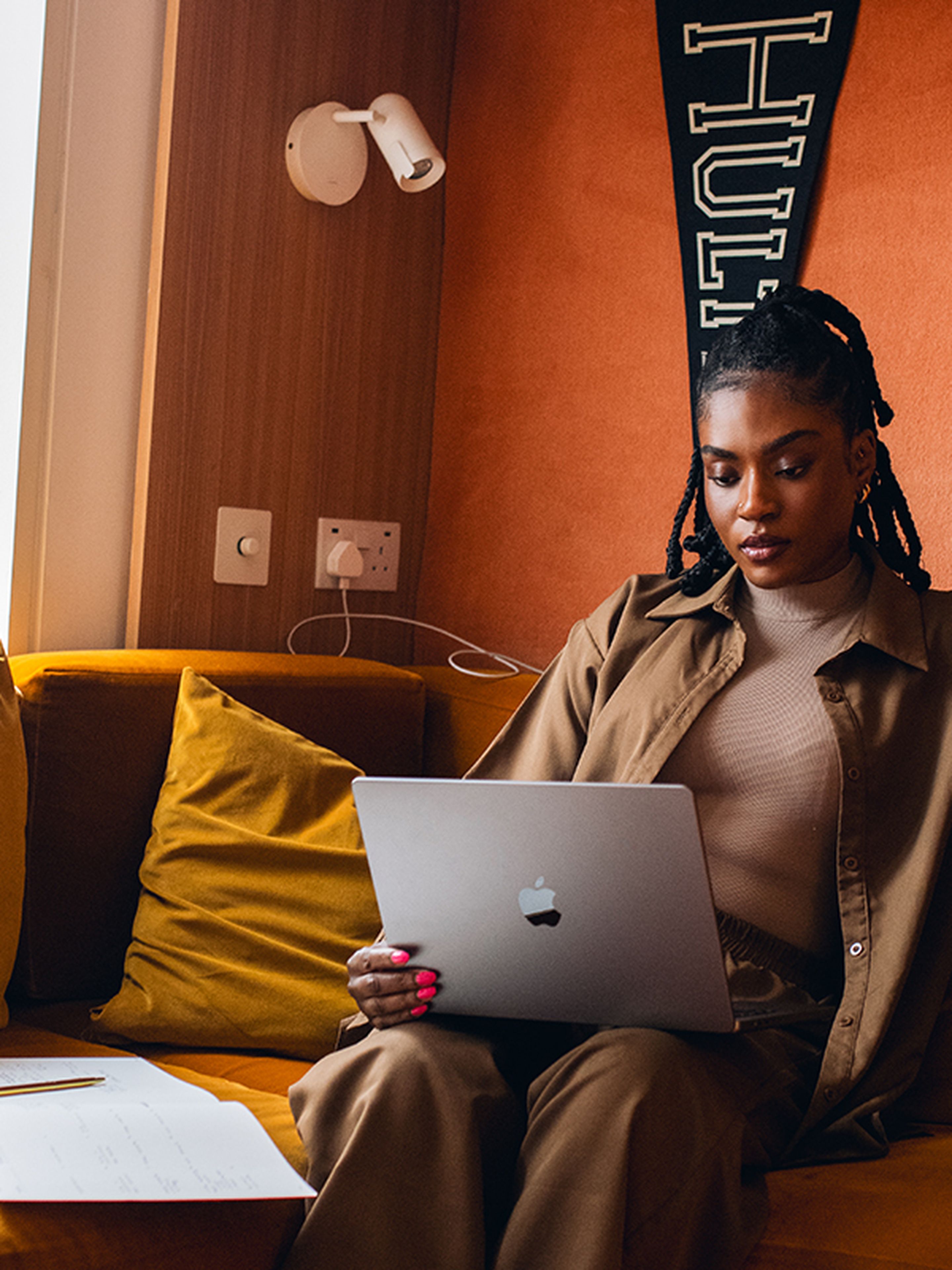 Woman sitting on a mustard sofa, using a laptop, with papers and a pen beside her in a cozy room with orange walls.