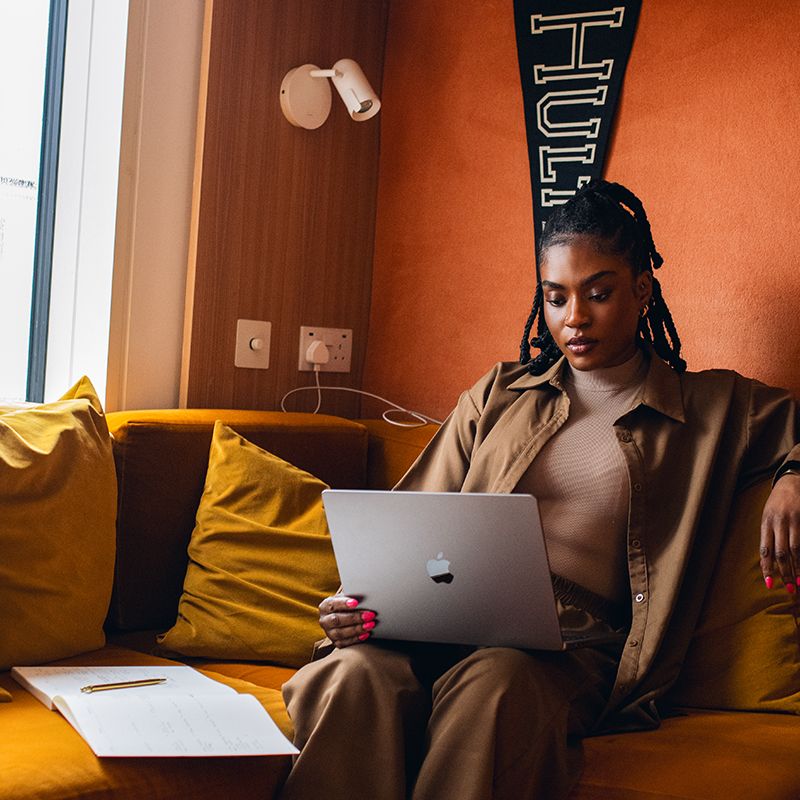 Woman sitting on a mustard sofa, using a laptop, with papers and a pen beside her in a cozy room with orange walls.
