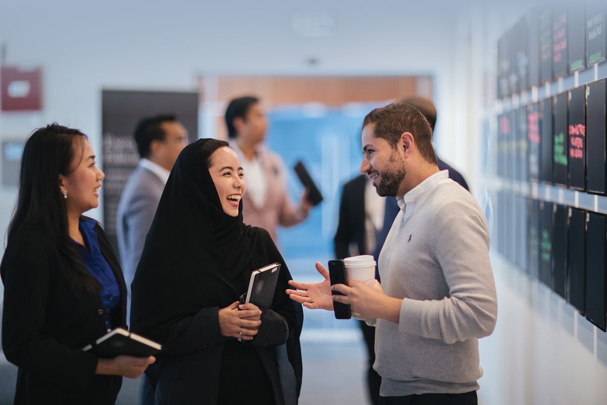 Three people conversing in a hallway, smiling and holding notebooks and a cup, with blurred figures in the background.