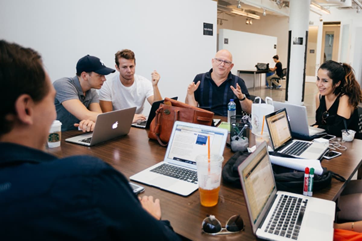 A group of people engaged in discussion around a table with laptops and drinks, in a modern office setting.