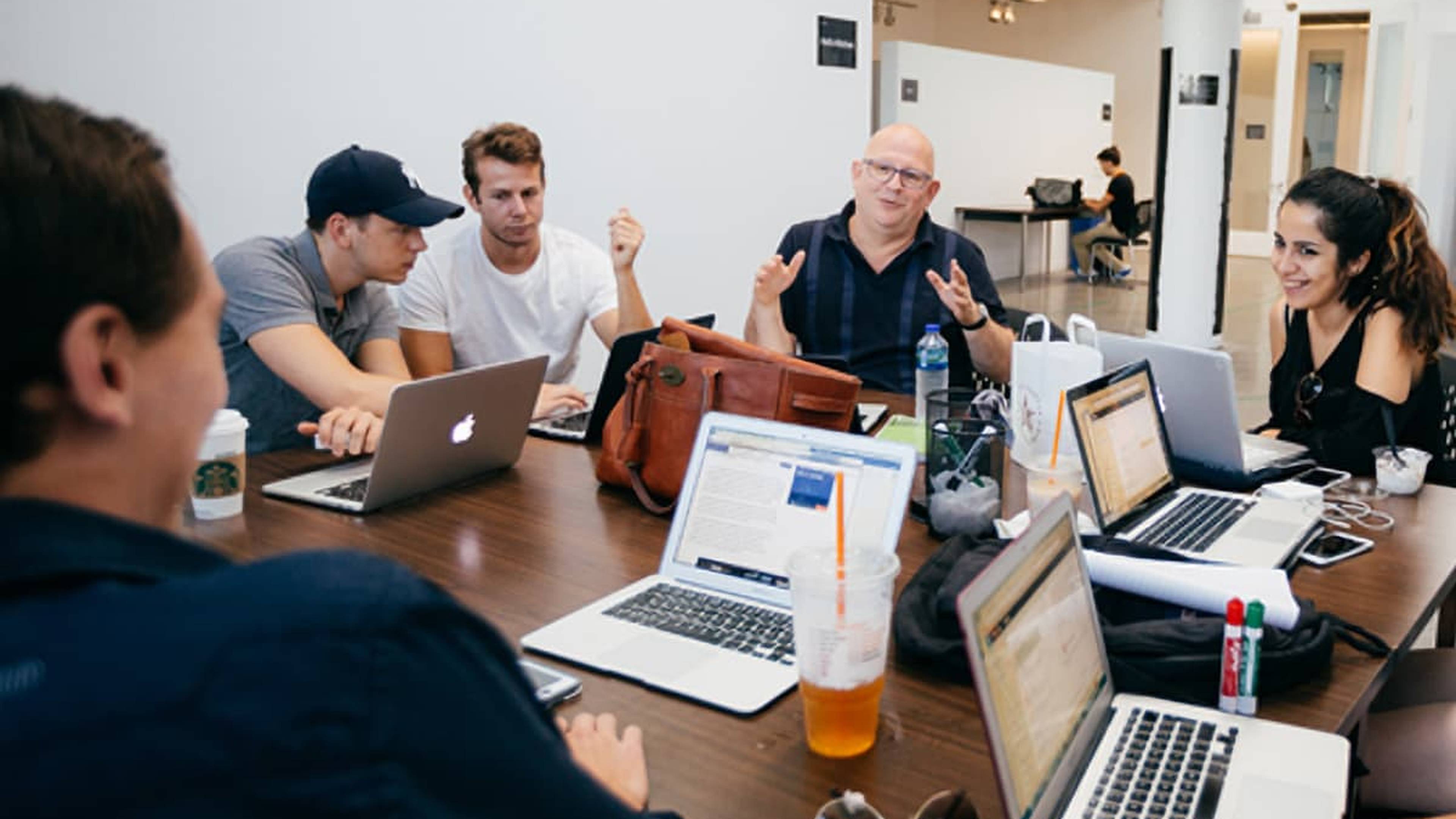 A group of people engaged in discussion around a table with laptops and drinks, in a modern office setting.