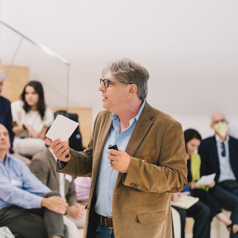 A man in a brown jacket speaks to an attentive audience in a tiered seating area, holding a microphone and notes.