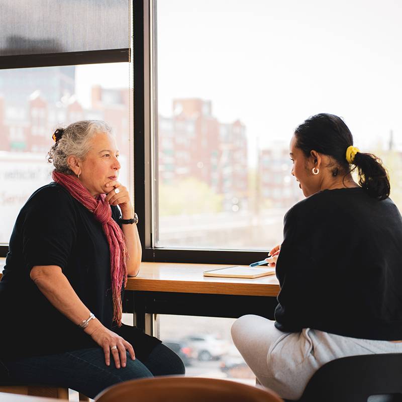 Two women sit at a table in front of large windows. One gestures while speaking, and the other listens and takes notes in a notebook.