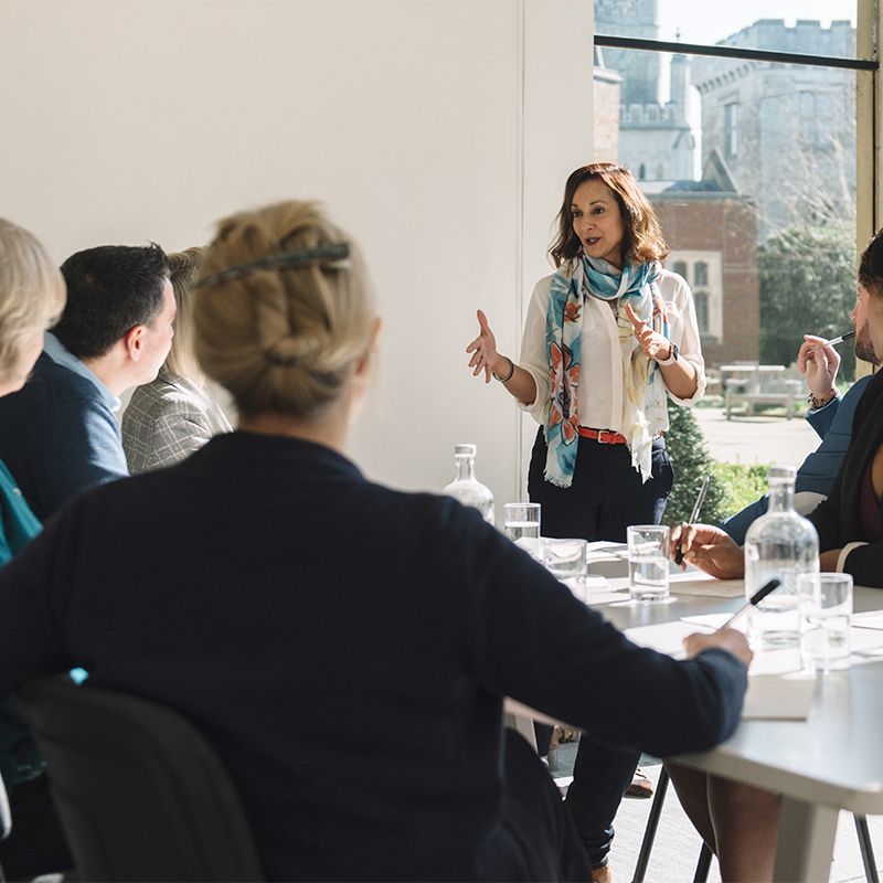 A woman is giving a presentation to a group seated around a table in a well-lit room with large windows overlooking a garden.