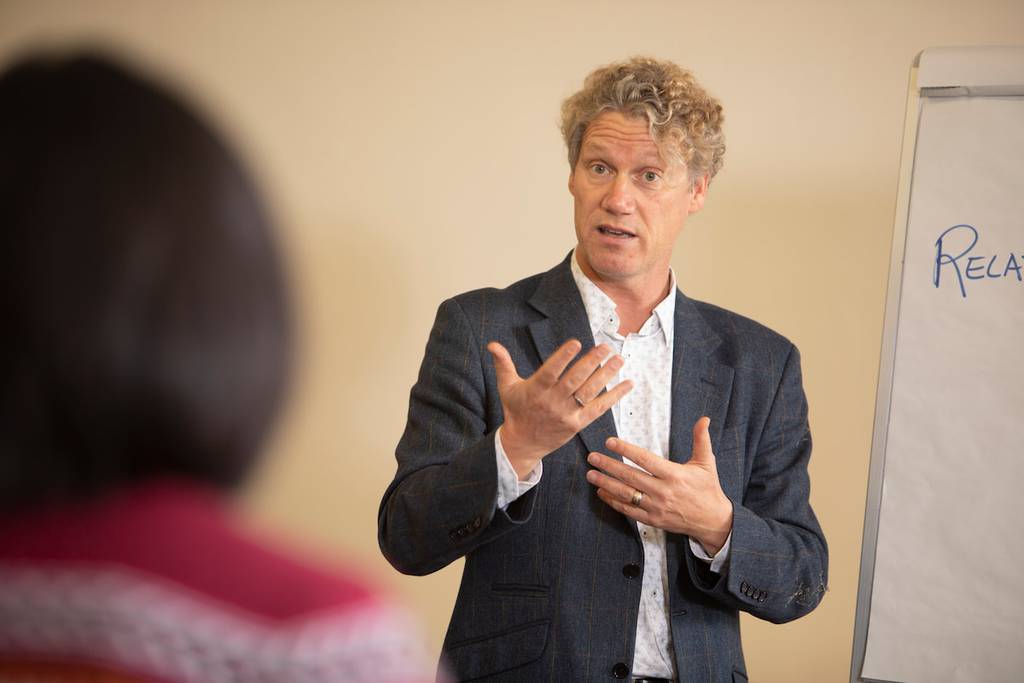 A man with curly hair gestures while speaking to an audience; a flipchart with partial text is visible beside him.