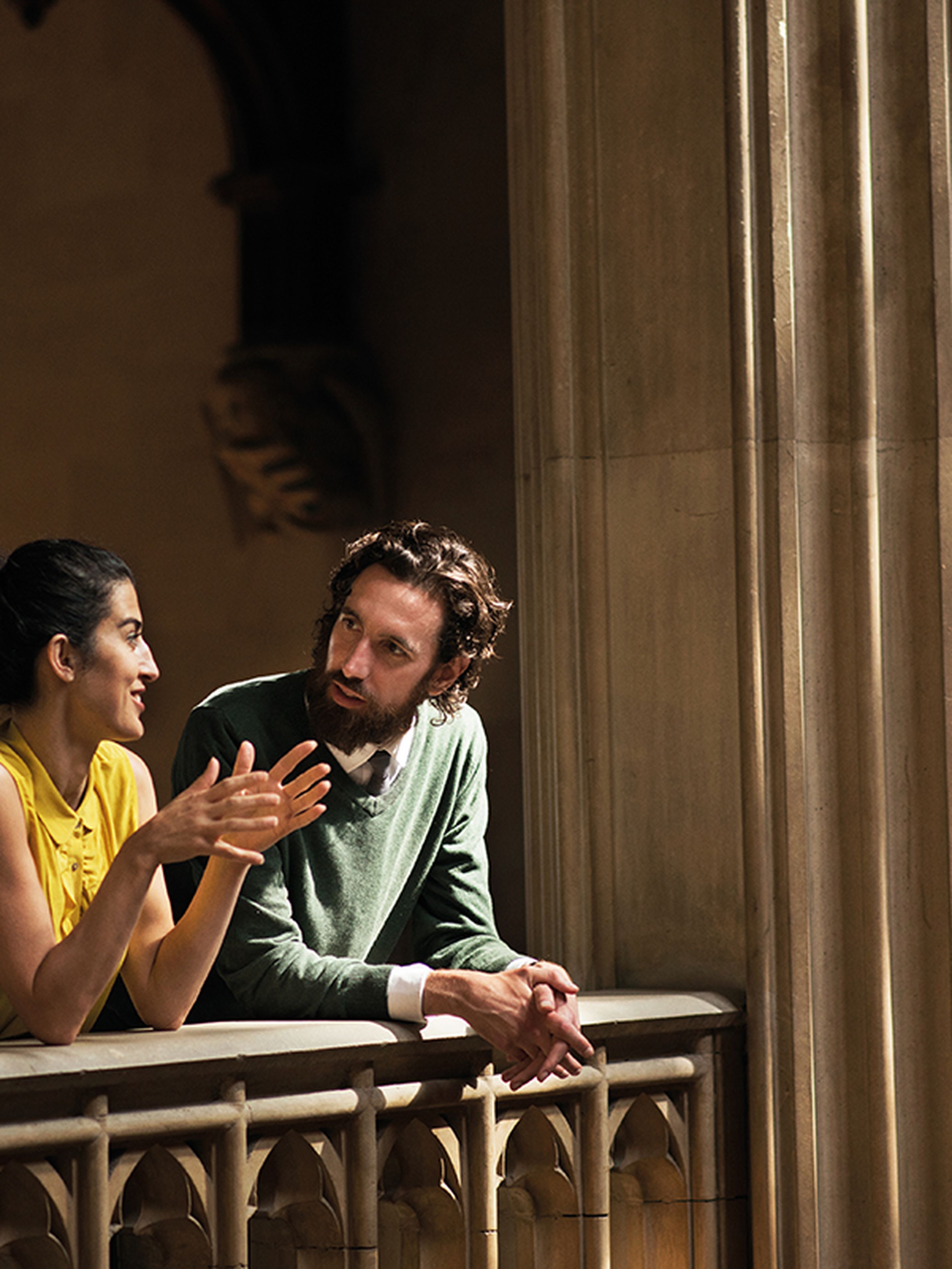 Two people are leaning on a stone railing, engaged in a lively conversation in a dimly lit setting with arches and columns.