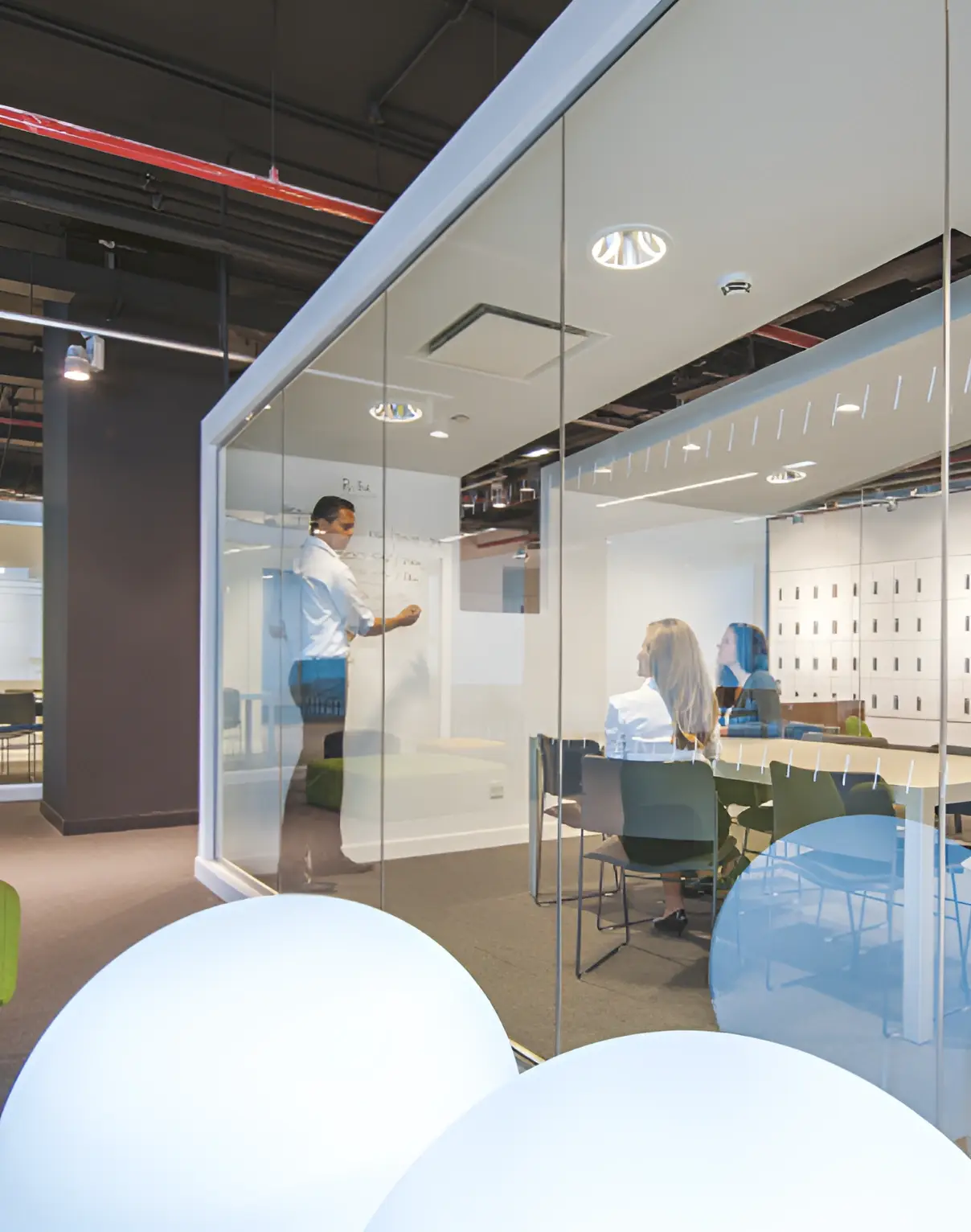 A person writes on a whiteboard while two people sit at a table inside a glass-walled office.