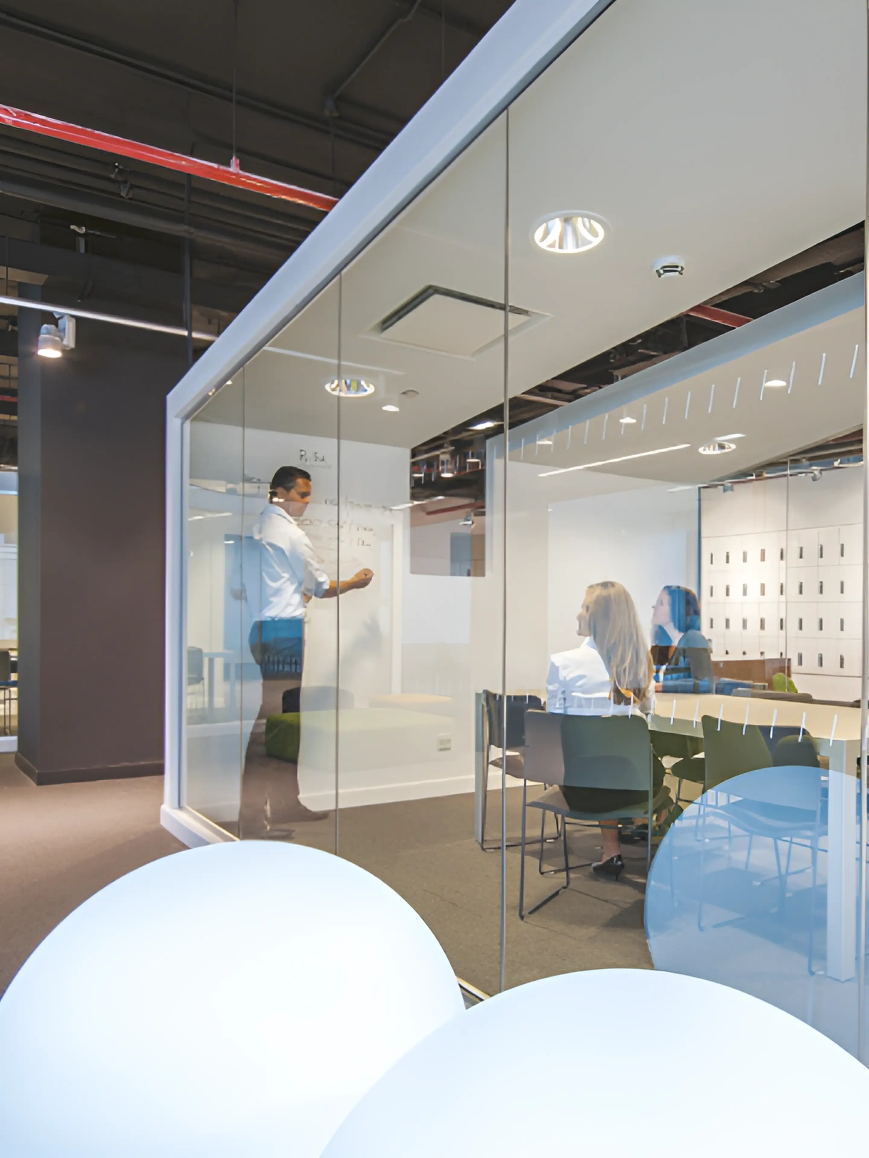 A person writes on a whiteboard while two people sit at a table inside a glass-walled office.