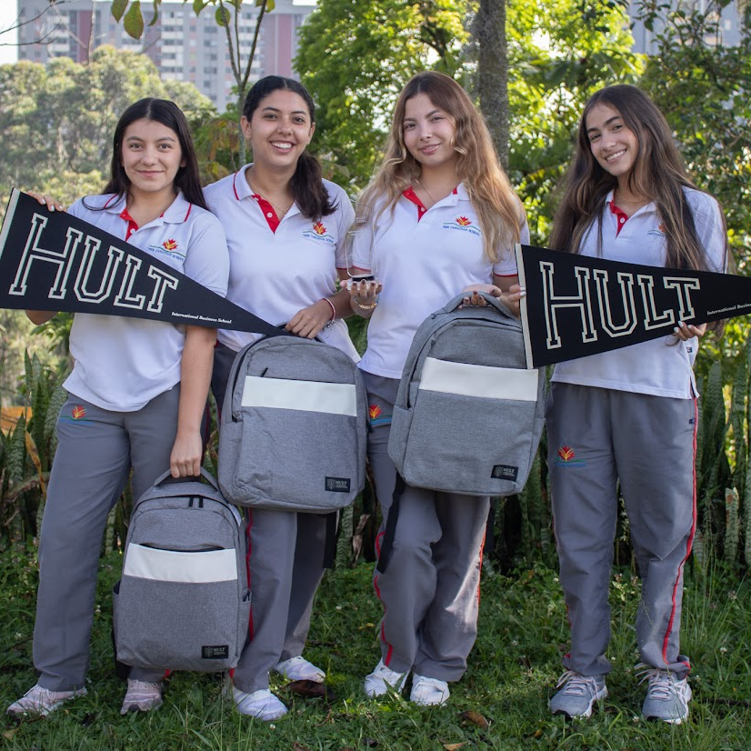 Four students in uniform stand outside holding "HULT" pennants and gray backpacks, smiling, with greenery and buildings in the background.