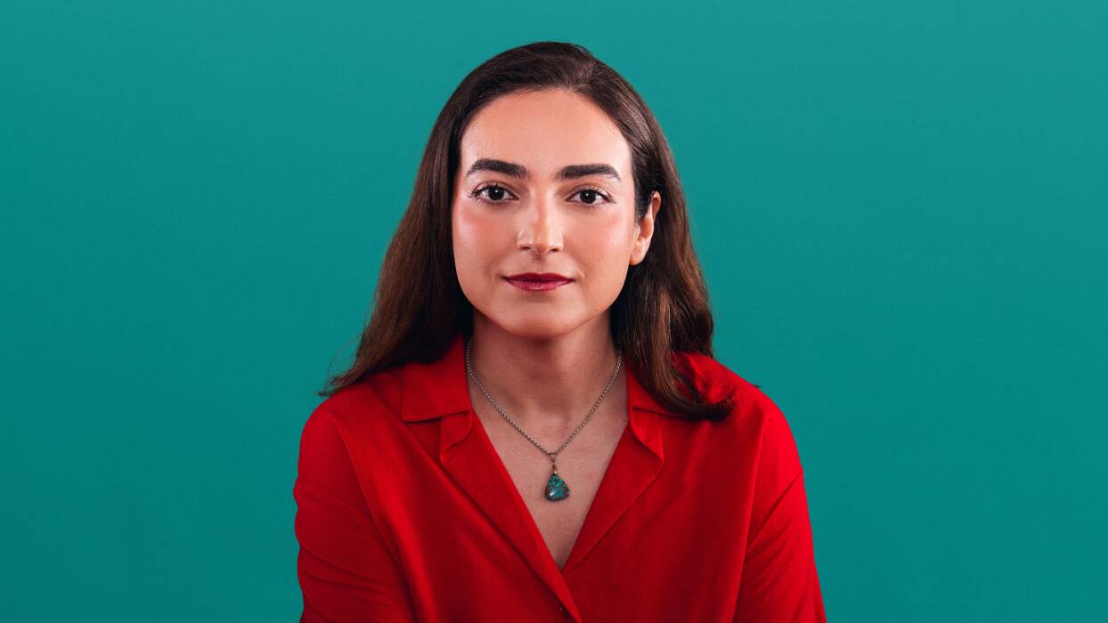 A woman in a red shirt and necklace poses against a teal background.