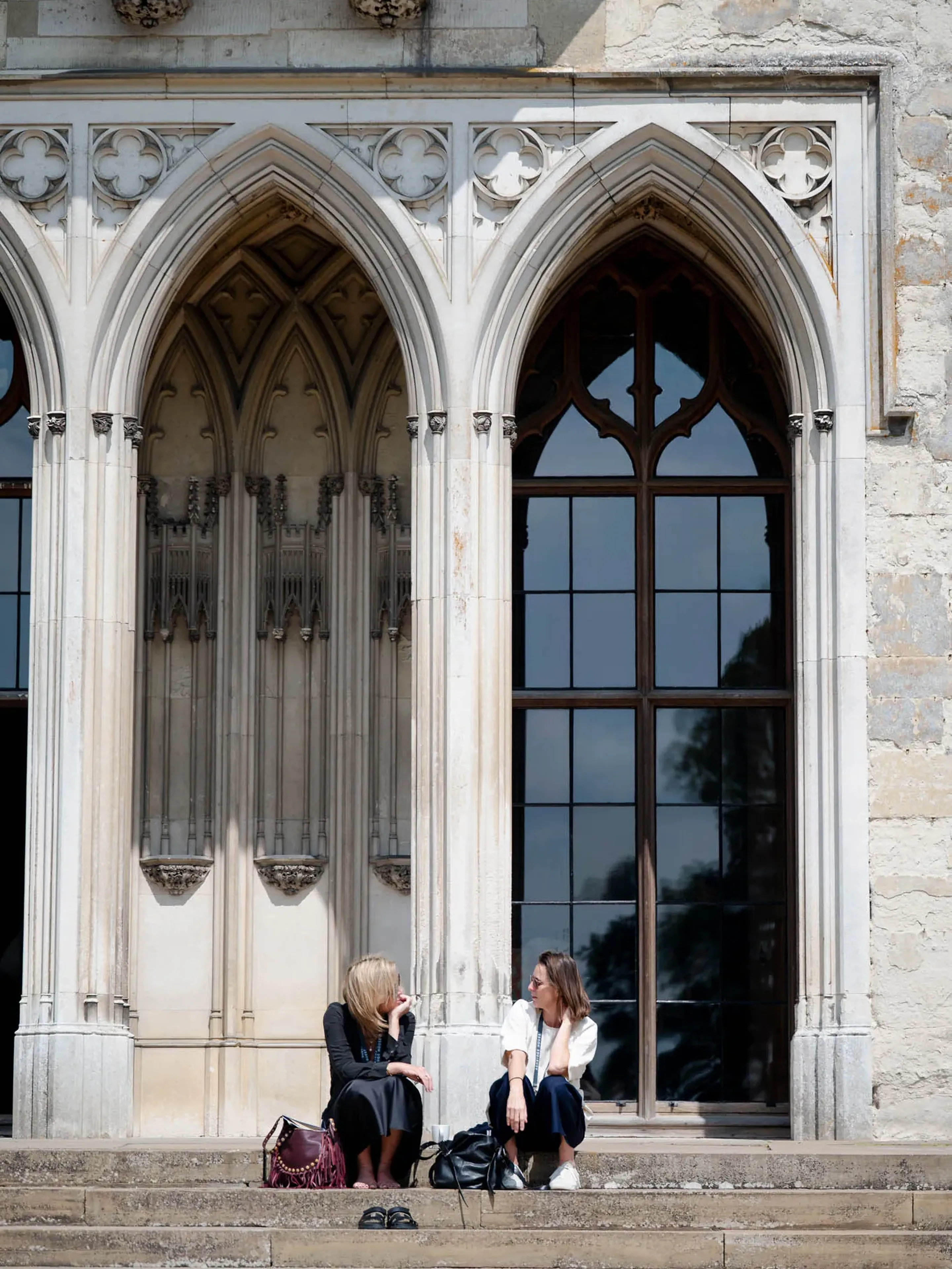 Two women sit and chat on steps outside a historic building with arched windows and ornate architecture.