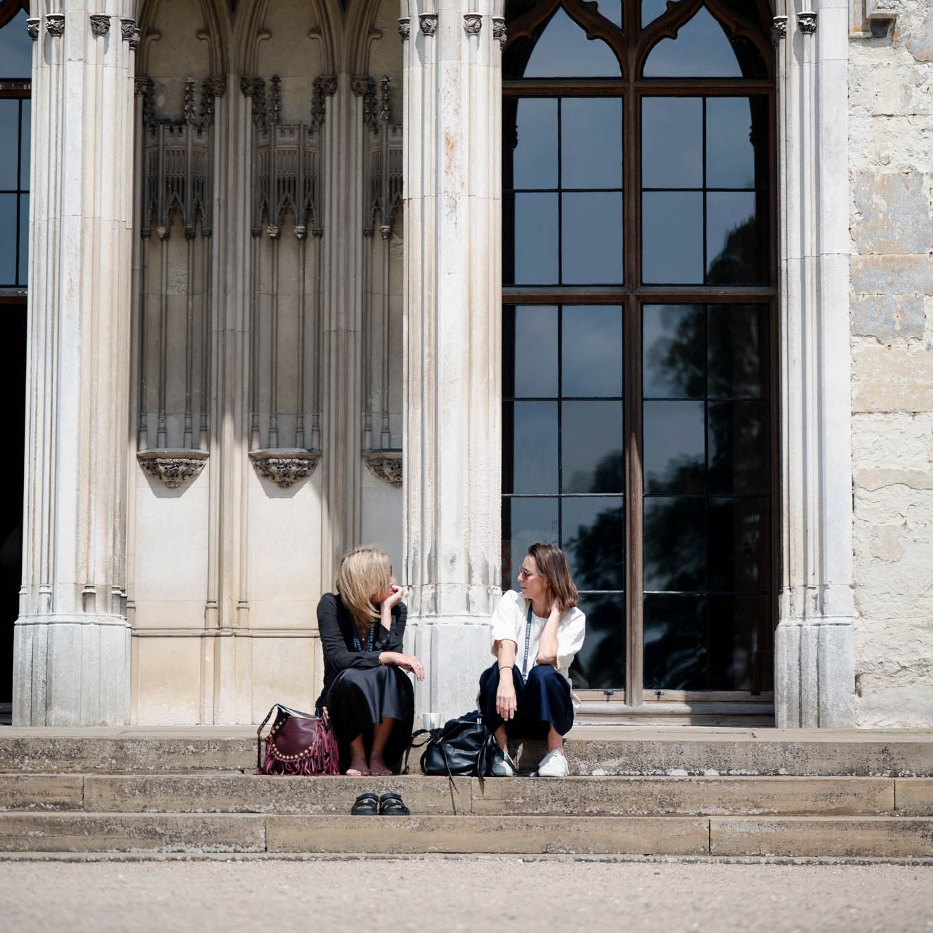Two women sitting on steps, talking in front of a Gothic arched entrance.