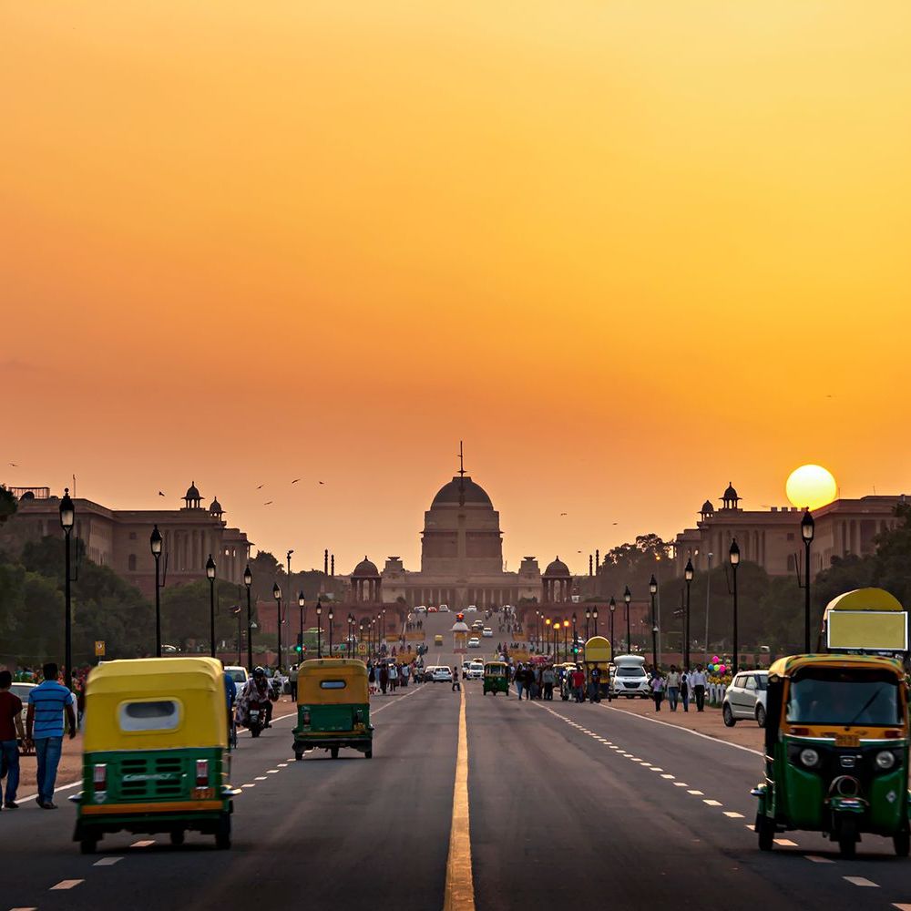 People and vehicles move along a wide road at sunset, with a domed building in the background.