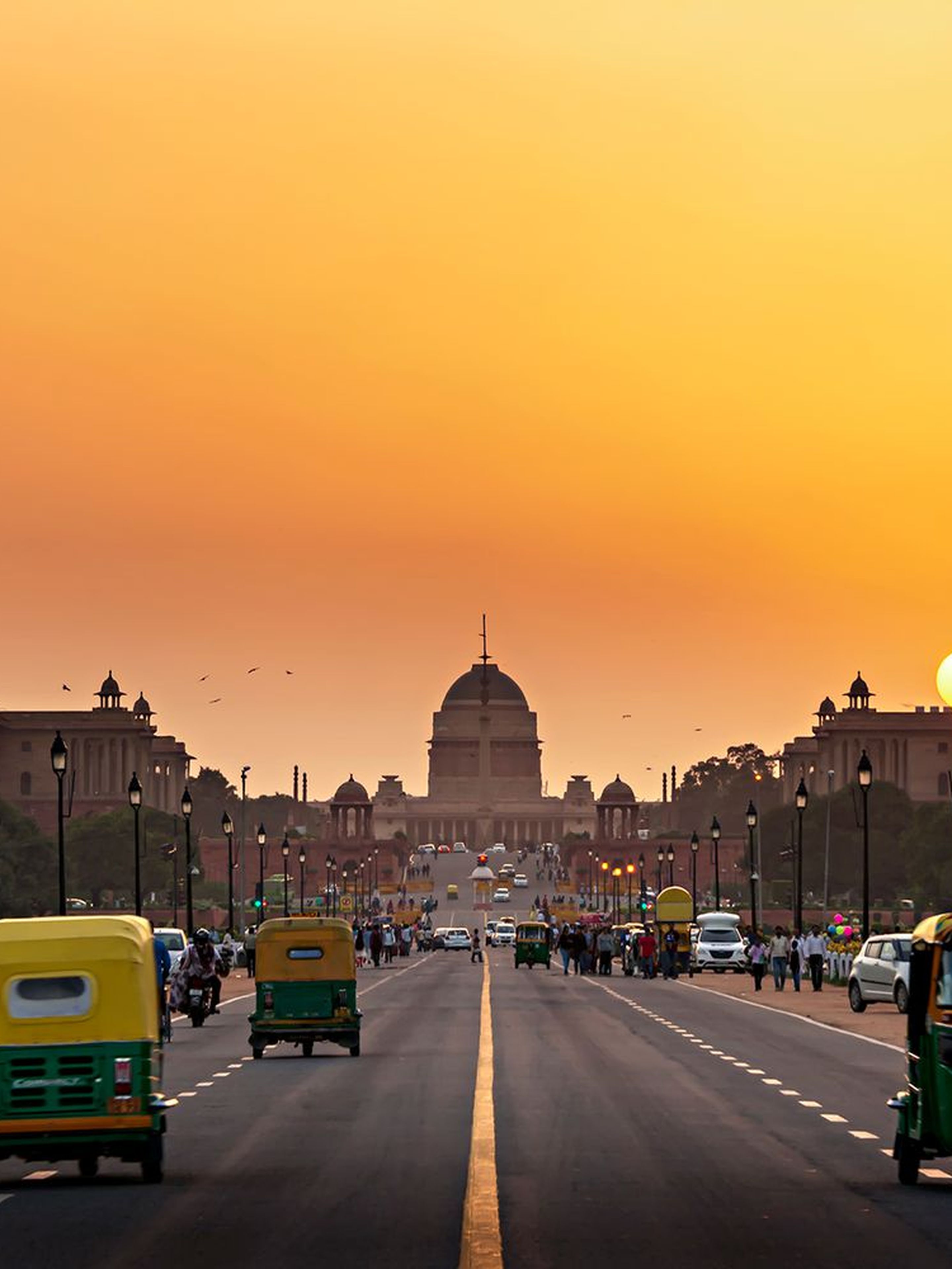 People and vehicles move along a wide road at sunset, with a domed building in the background.