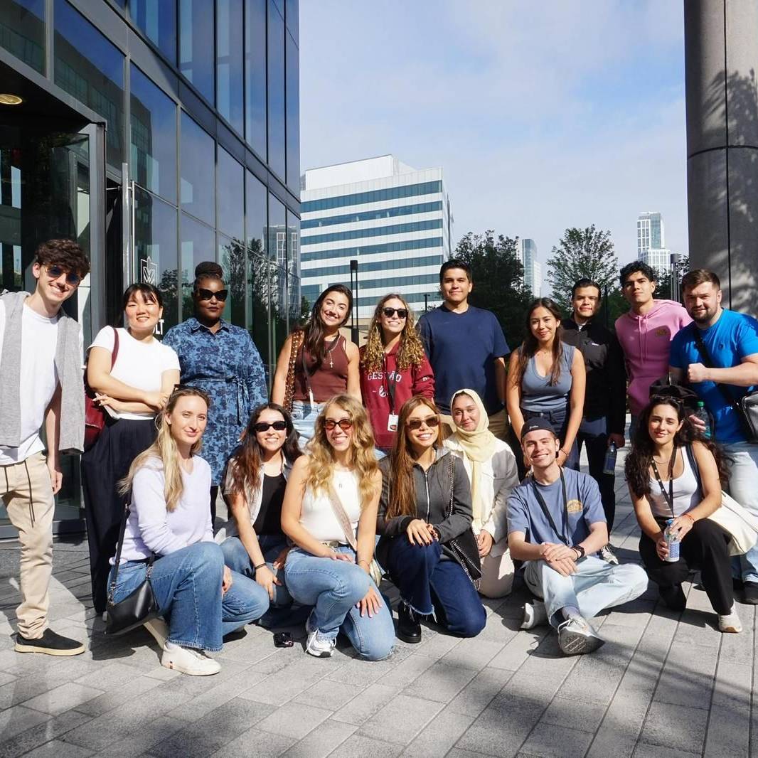 A diverse group of people smiling and posing together outside a modern building on a sunny day.