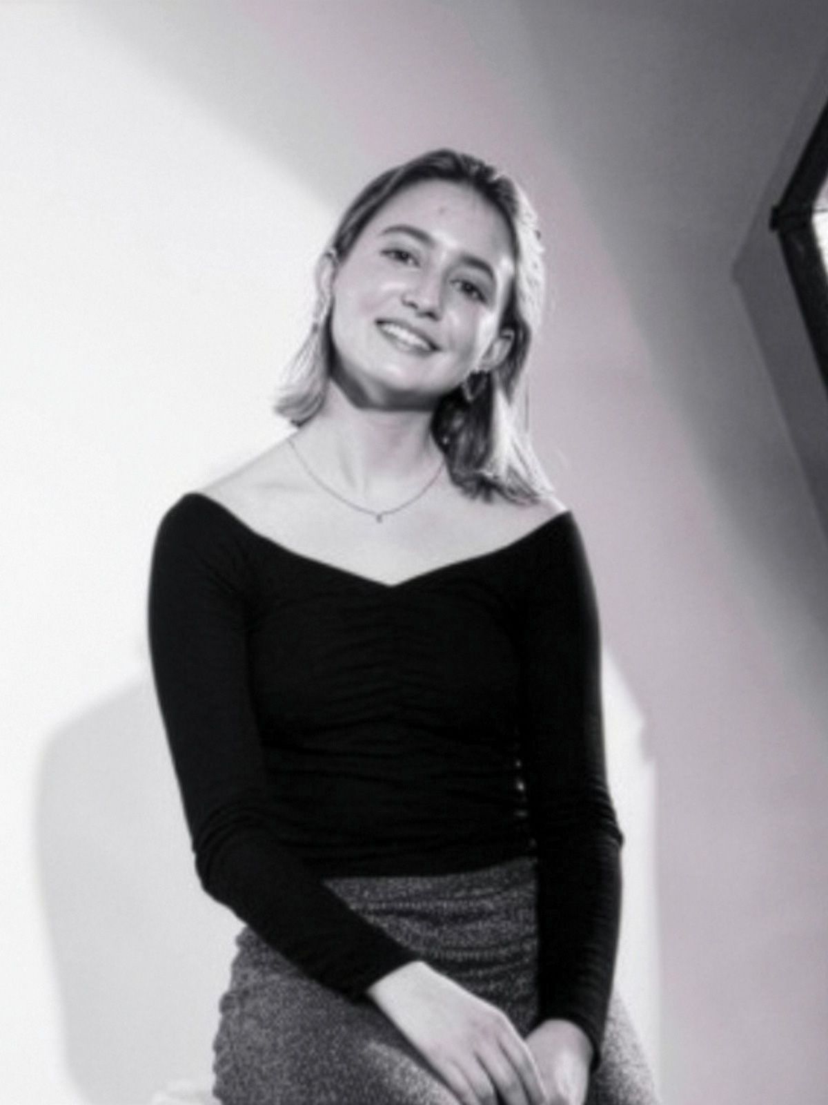 Woman seated, smiling in a studio setting. She wears a black top, skirt, and a necklace. Soft lighting from a nearby diffuser creates gentle shadows.