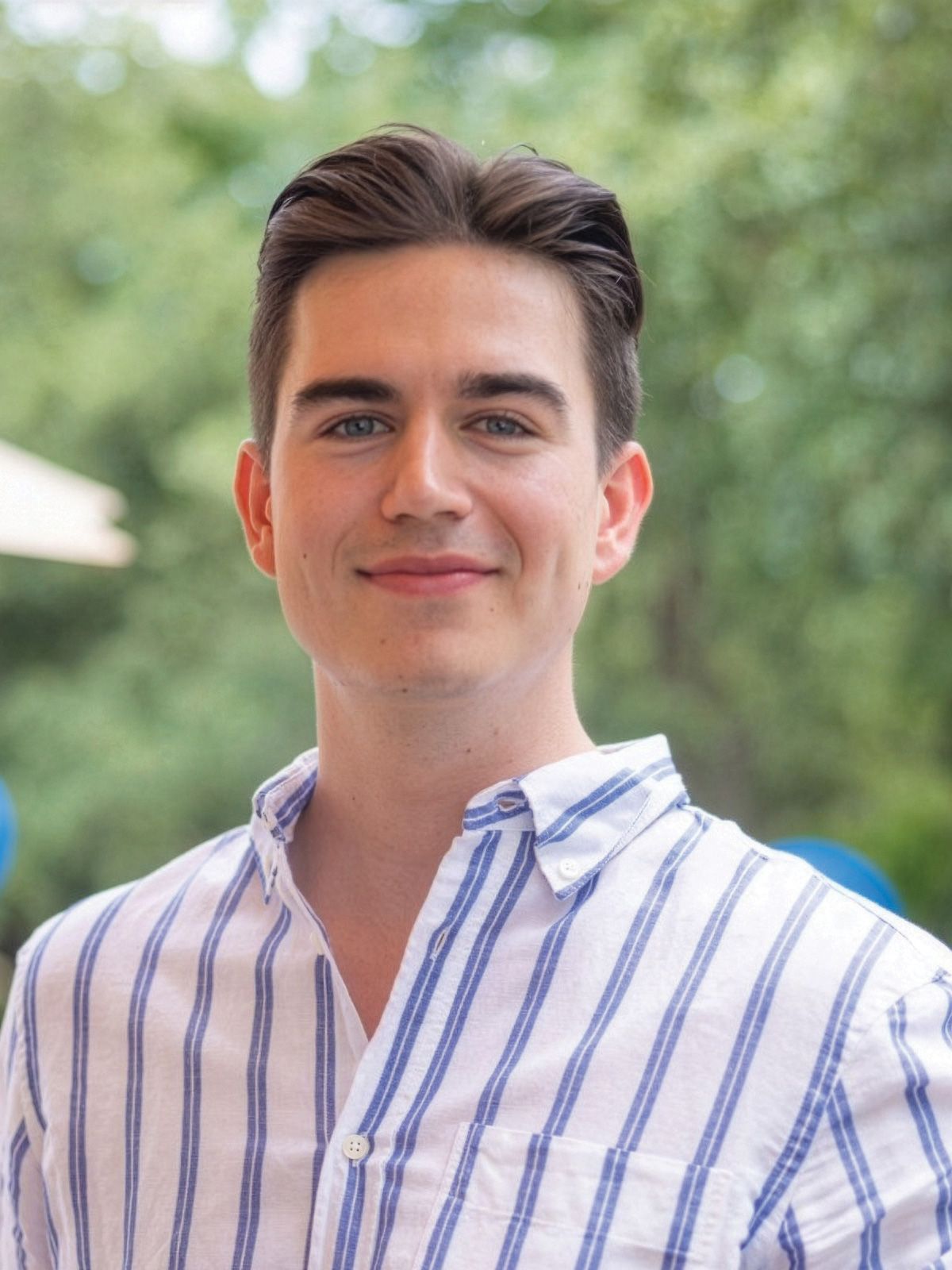 Smiling individual in a striped shirt standing outdoors with blurred greenery and blue balloons in the background.