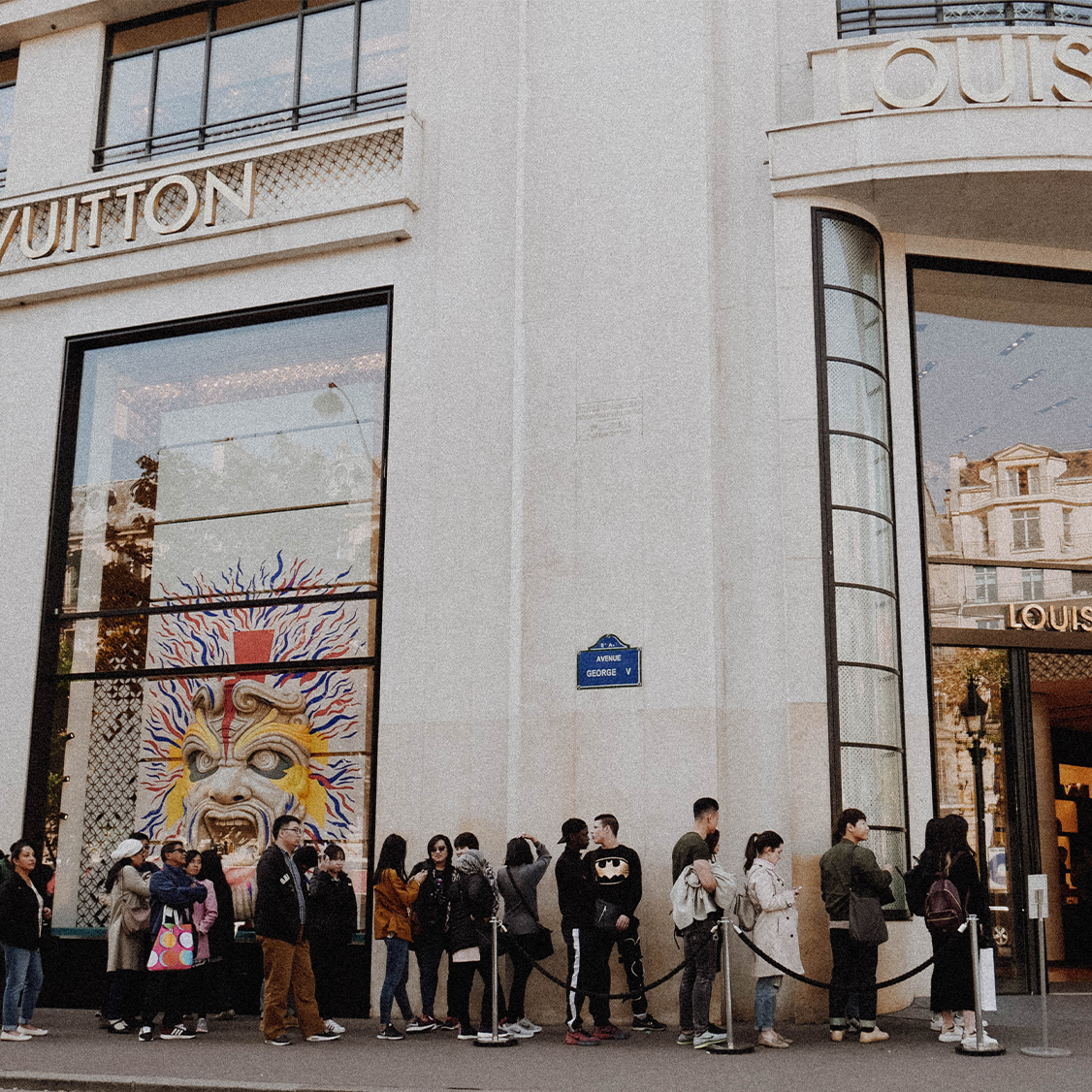 People line up outside a Louis Vuitton store with large windows and a colorful mural.