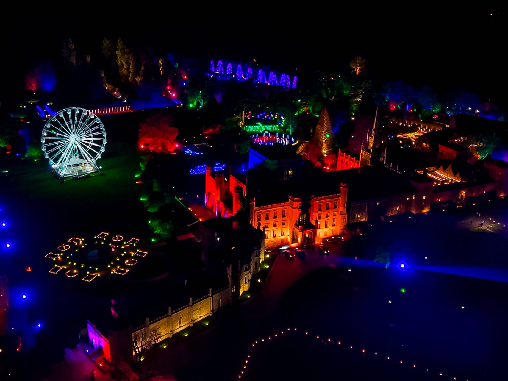 Aerial view of a festive nighttime scene with vibrant lights, featuring a Ferris wheel and illuminated buildings in a park setting.