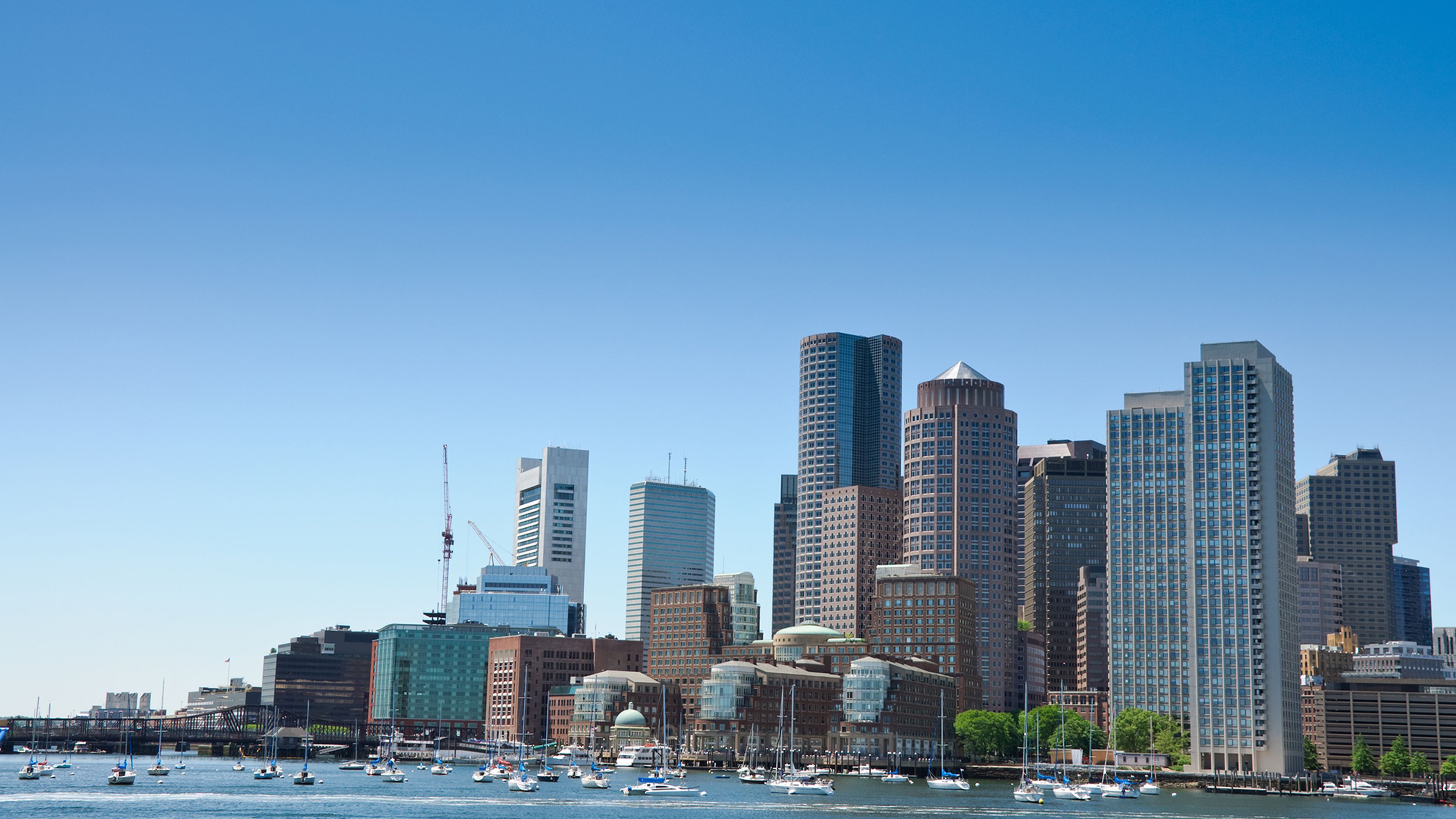 Boston skyline with skyscrapers and boats on the water under a clear blue sky.