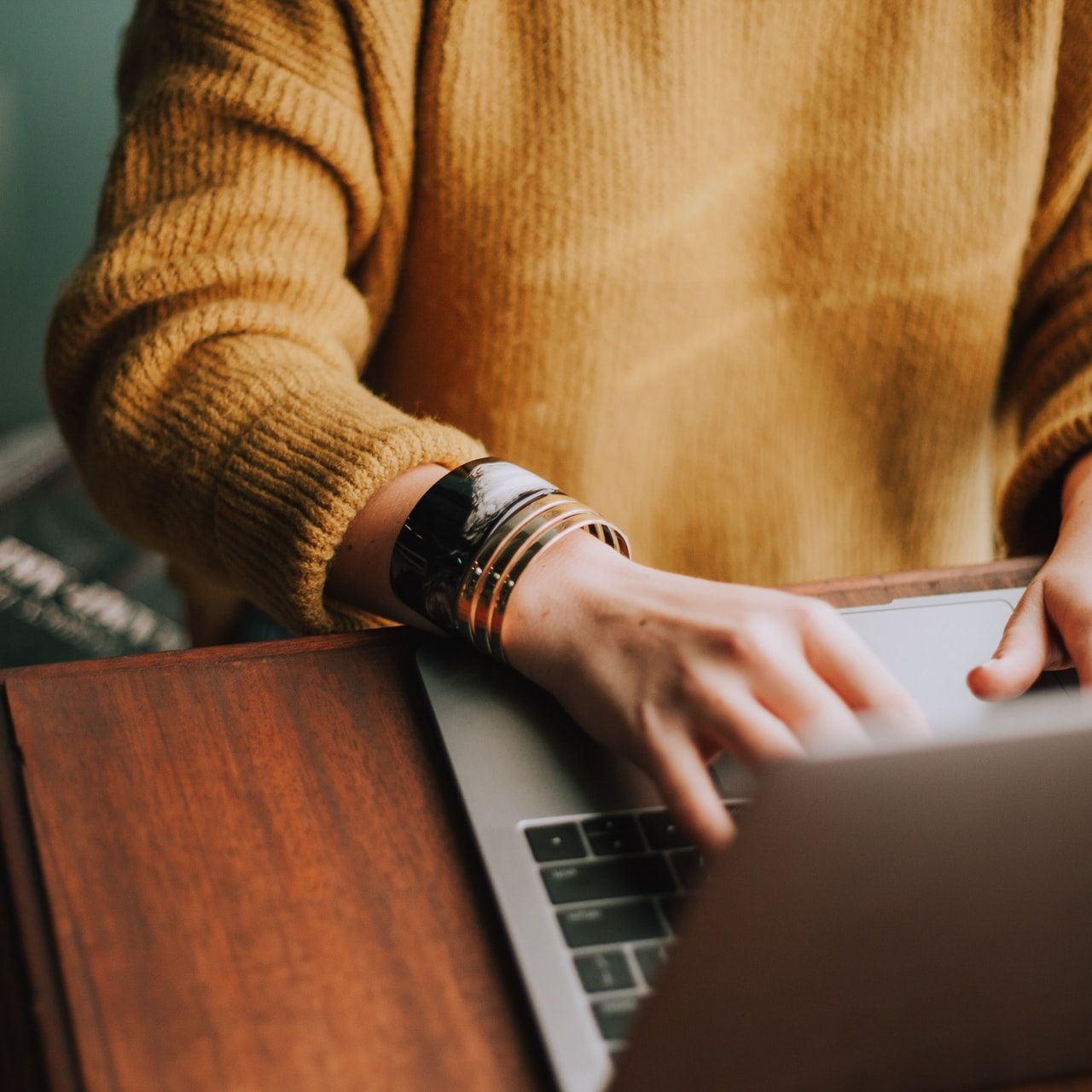 Person in a yellow sweater typing on a laptop at a wooden desk.