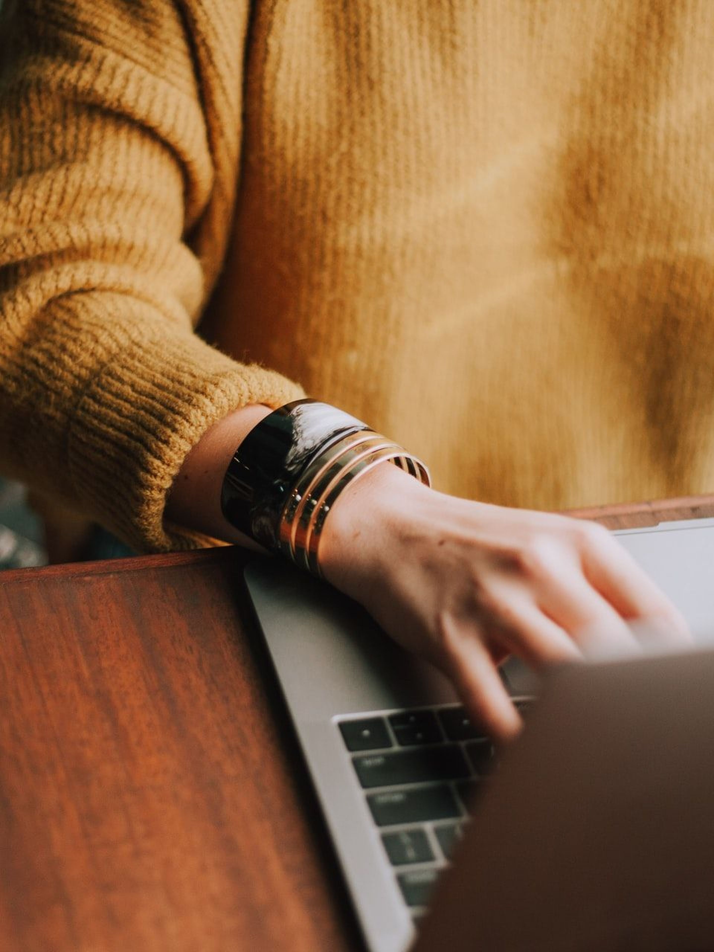 Person in a yellow sweater typing on a laptop at a wooden desk.