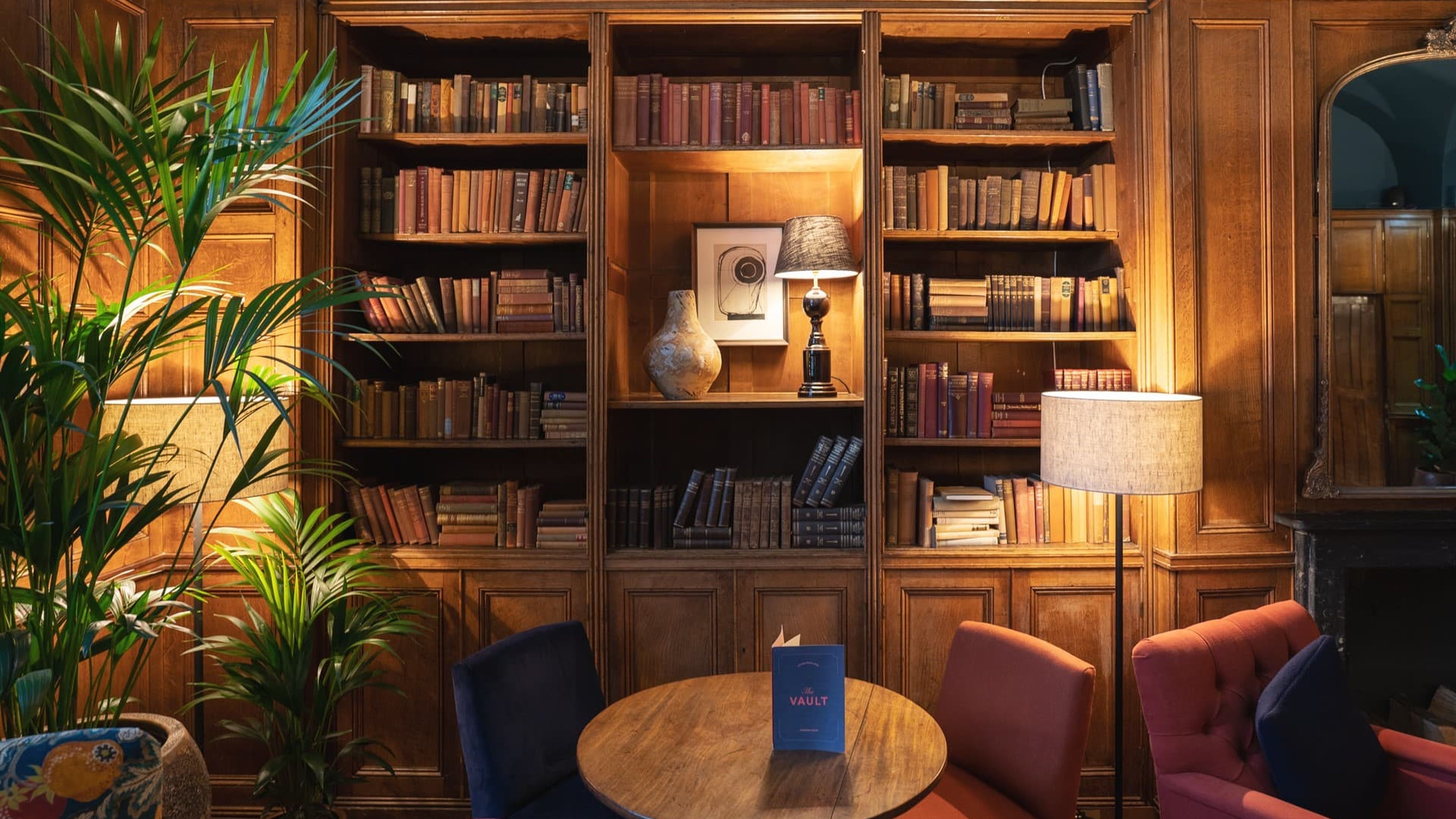 Cozy library corner with wooden bookshelves, books, warm lighting, and a small round table surrounded by colorful upholstered chairs.