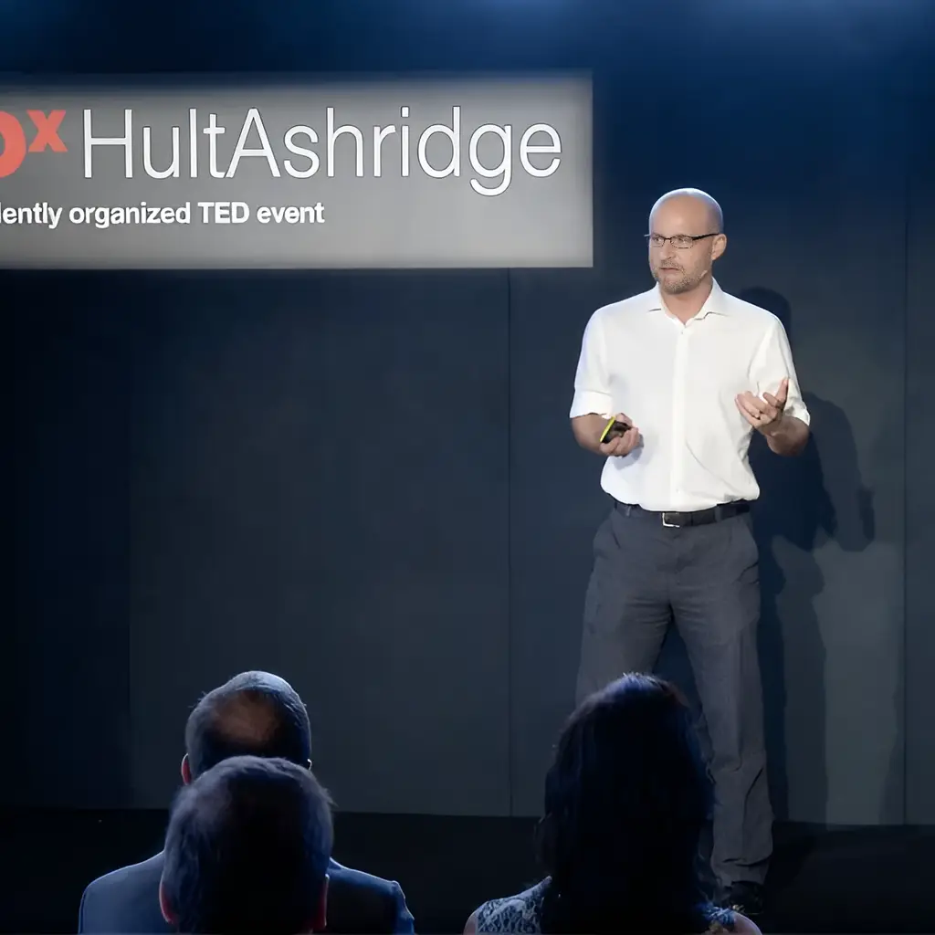 A speaker in a white shirt presents at TEDxHultAshridge event, facing an audience.
