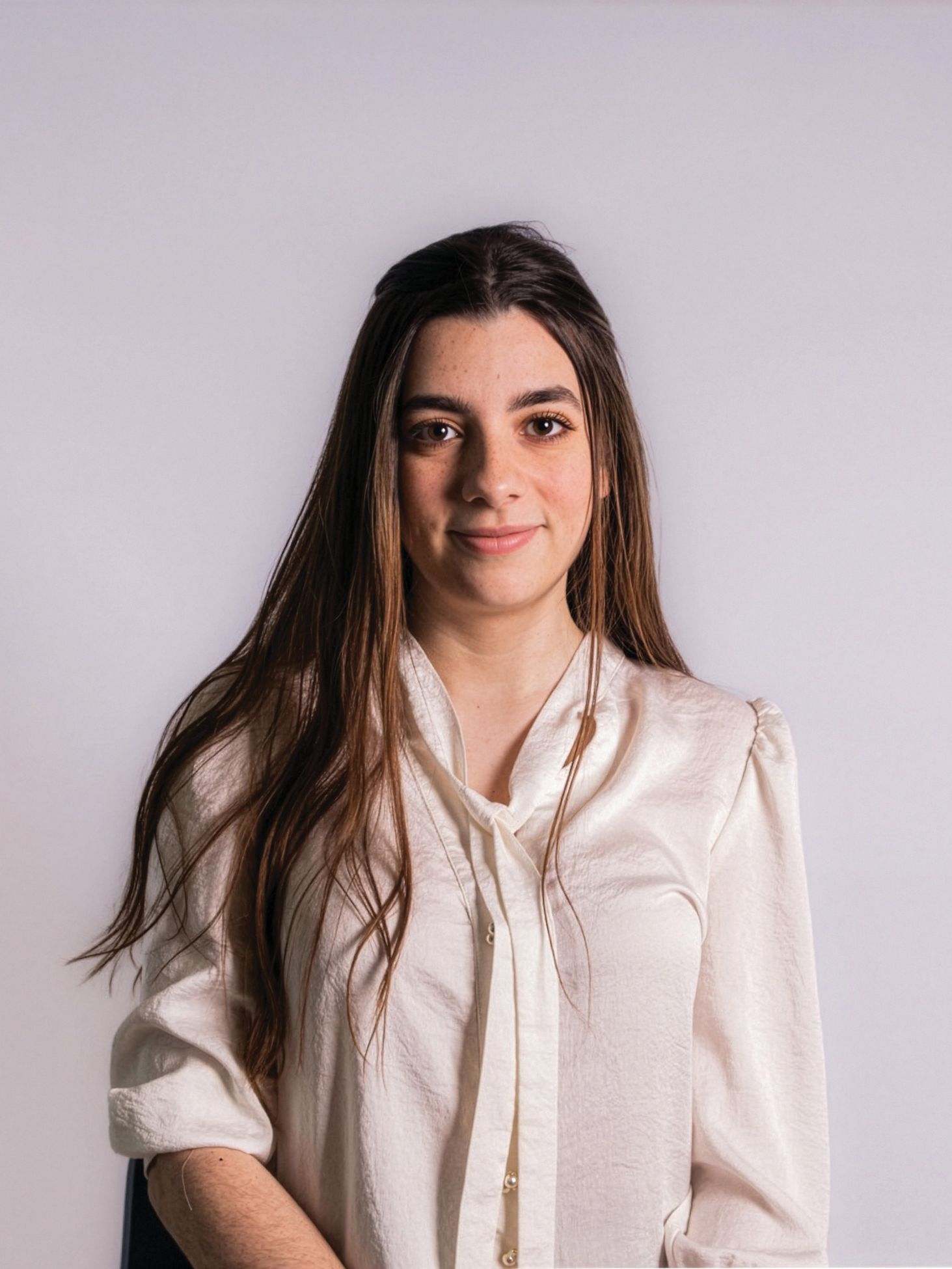 Young woman with long hair in a white blouse, standing against a plain light background, looking at the camera.