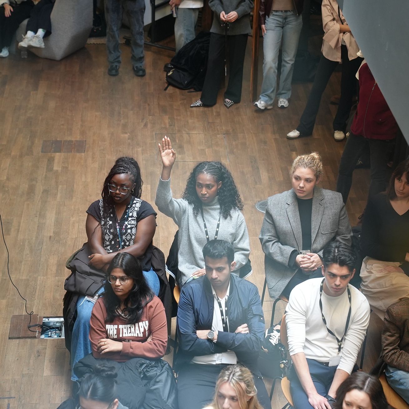 A person raises their hand during a seated gathering in a room with a wooden floor.