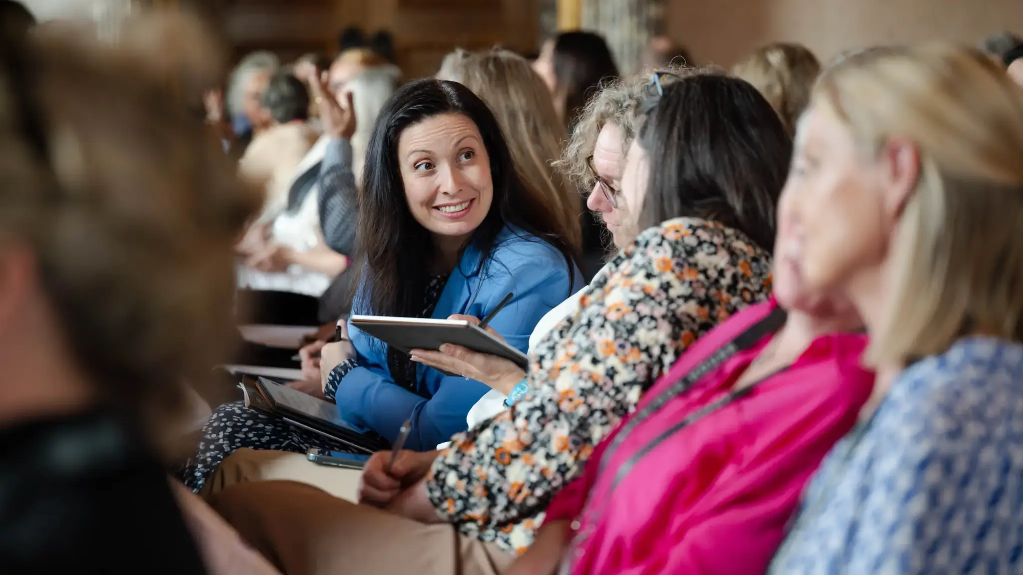 A group of people seated in a conference room, engaged in conversation. One person in a blue jacket smiles while holding a notebook.