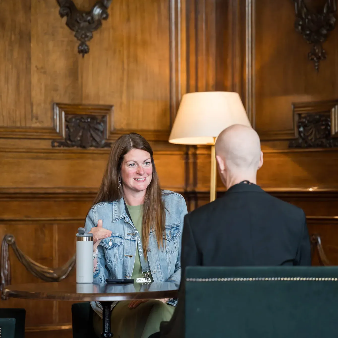 A woman in a denim jacket smiles while talking to a person in a black coat, seated at a table in a room with wooden paneling and a lamp.