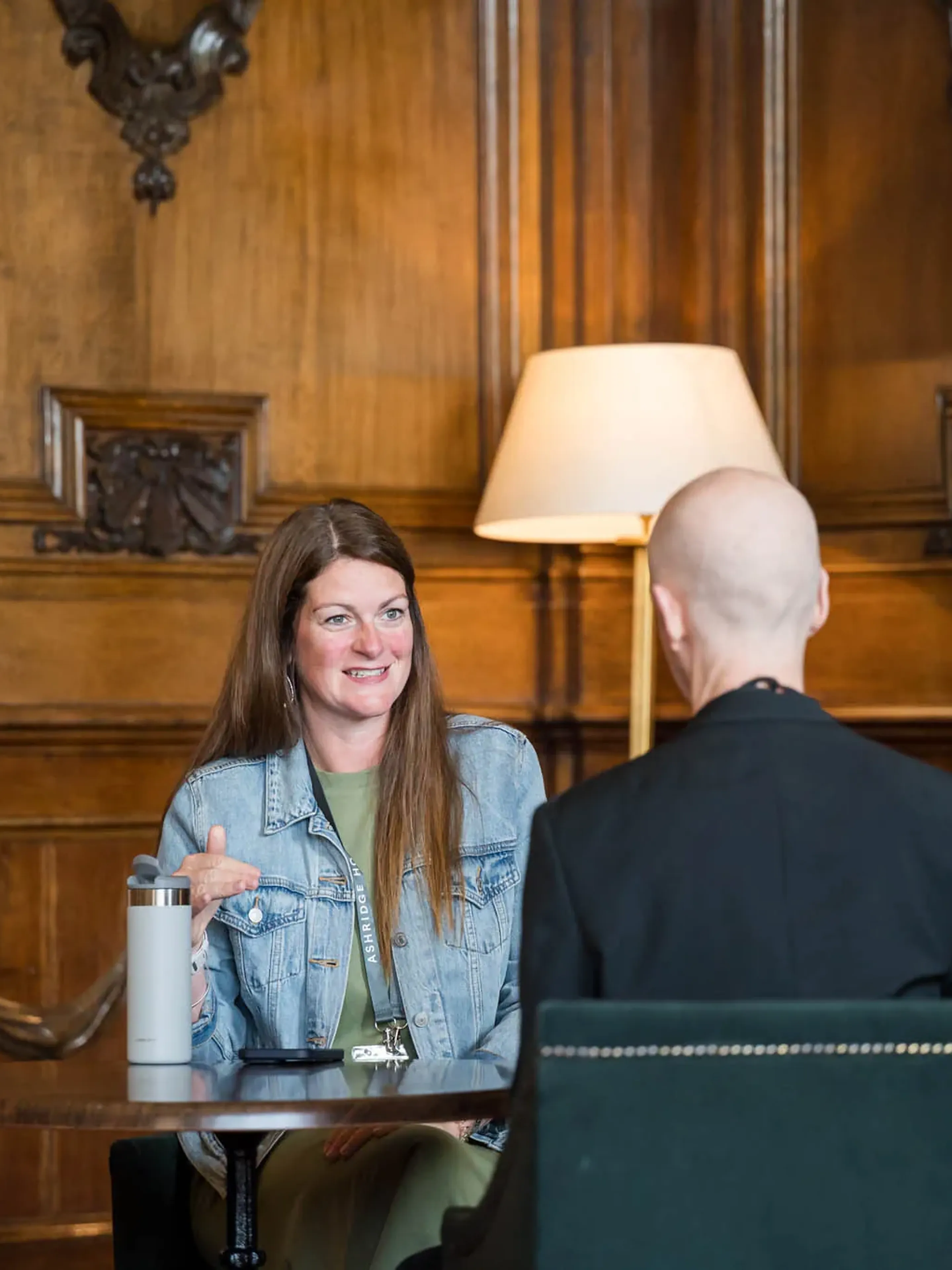 A woman in a denim jacket smiles while talking to a person in a black coat, seated at a table in a room with wooden paneling and a lamp.