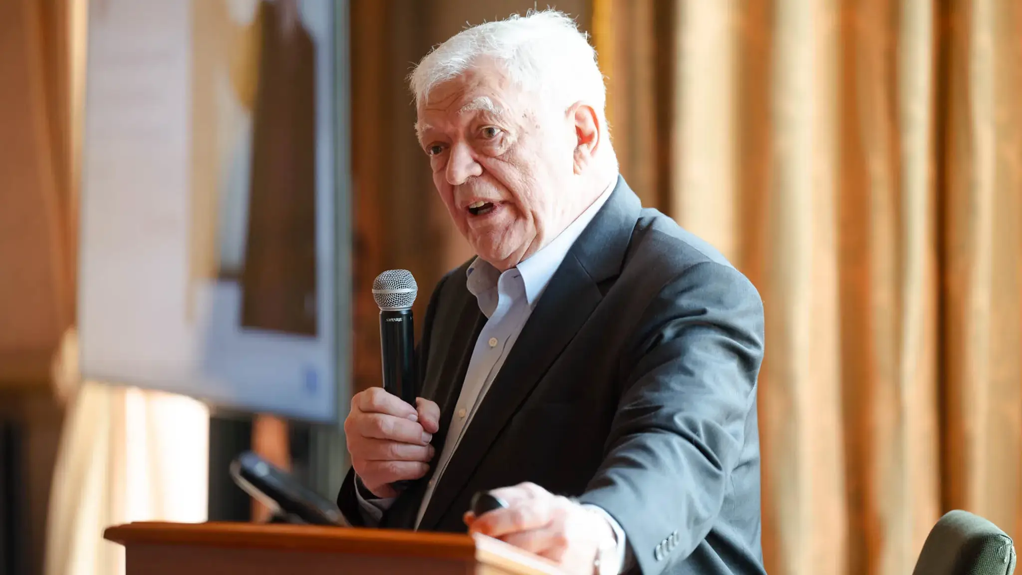 Elderly man in a suit speaking into a microphone at a podium inside a room with curtains and a blurry background.